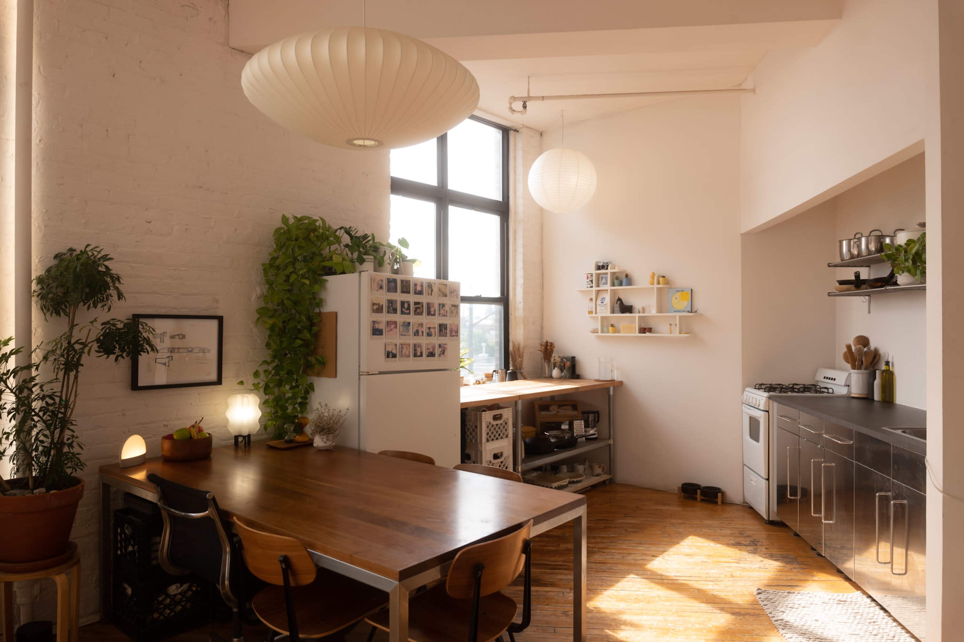 A bright kitchen area features a wooden dining table, a refrigerator adorned with photos, and a section of open shelving against a wall.