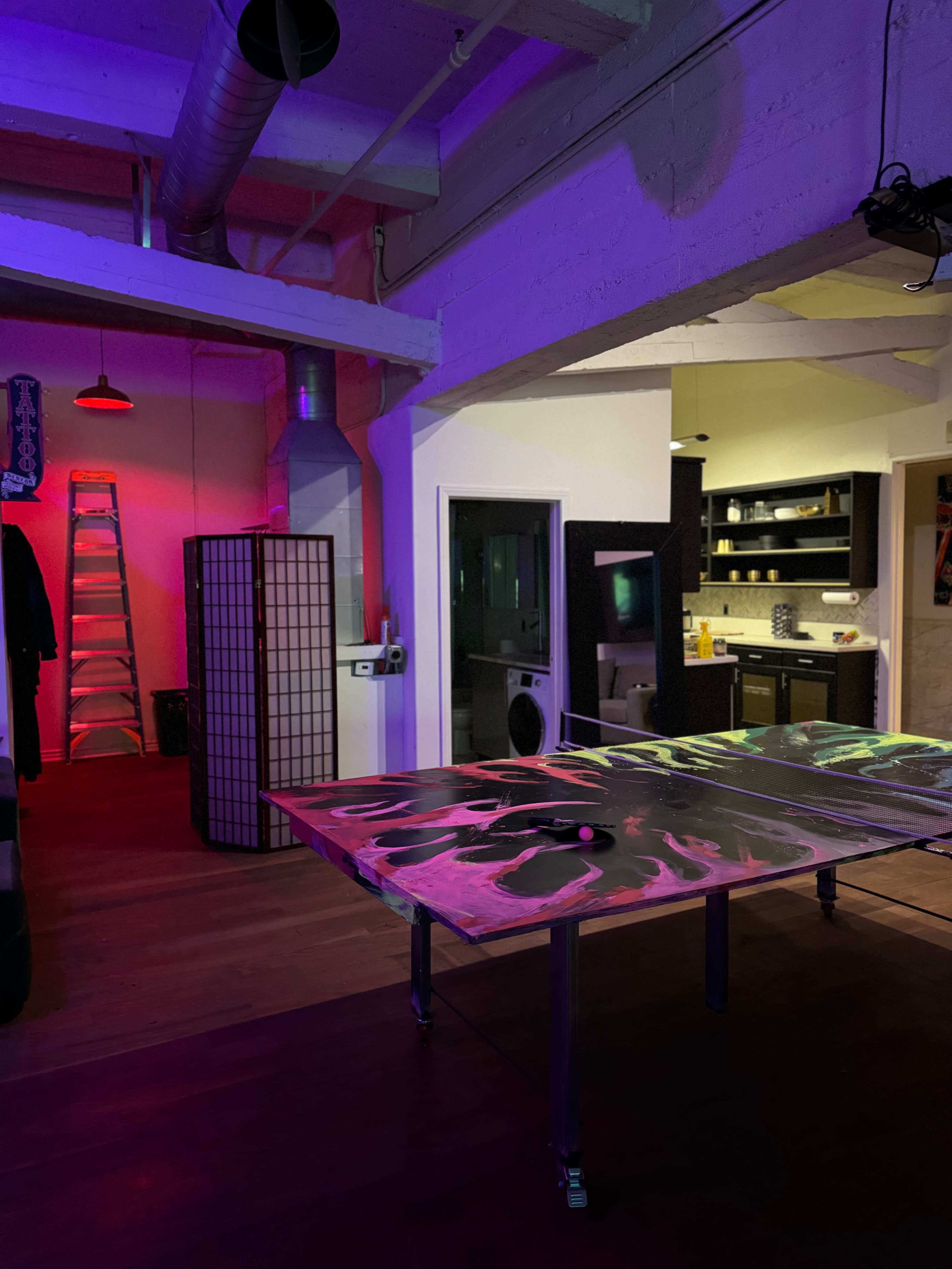 The image shows a vibrant indoor space featuring a ping pong table, a ladder, and a kitchen area illuminated by colored lights.