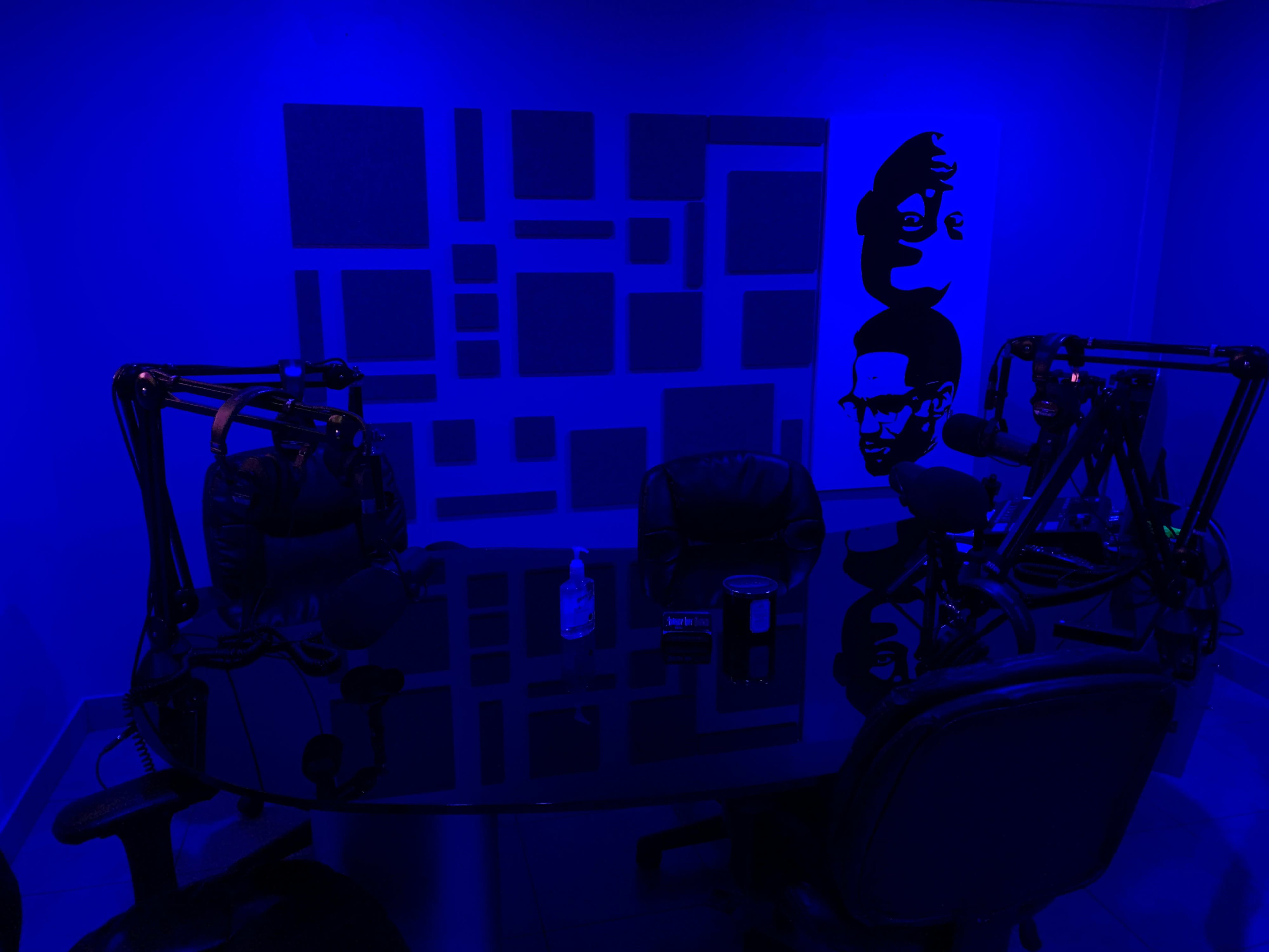 A dimly lit podcast studio with a black table, two chairs, and microphone stands against a blue wall featuring abstract designs and a face illustration.