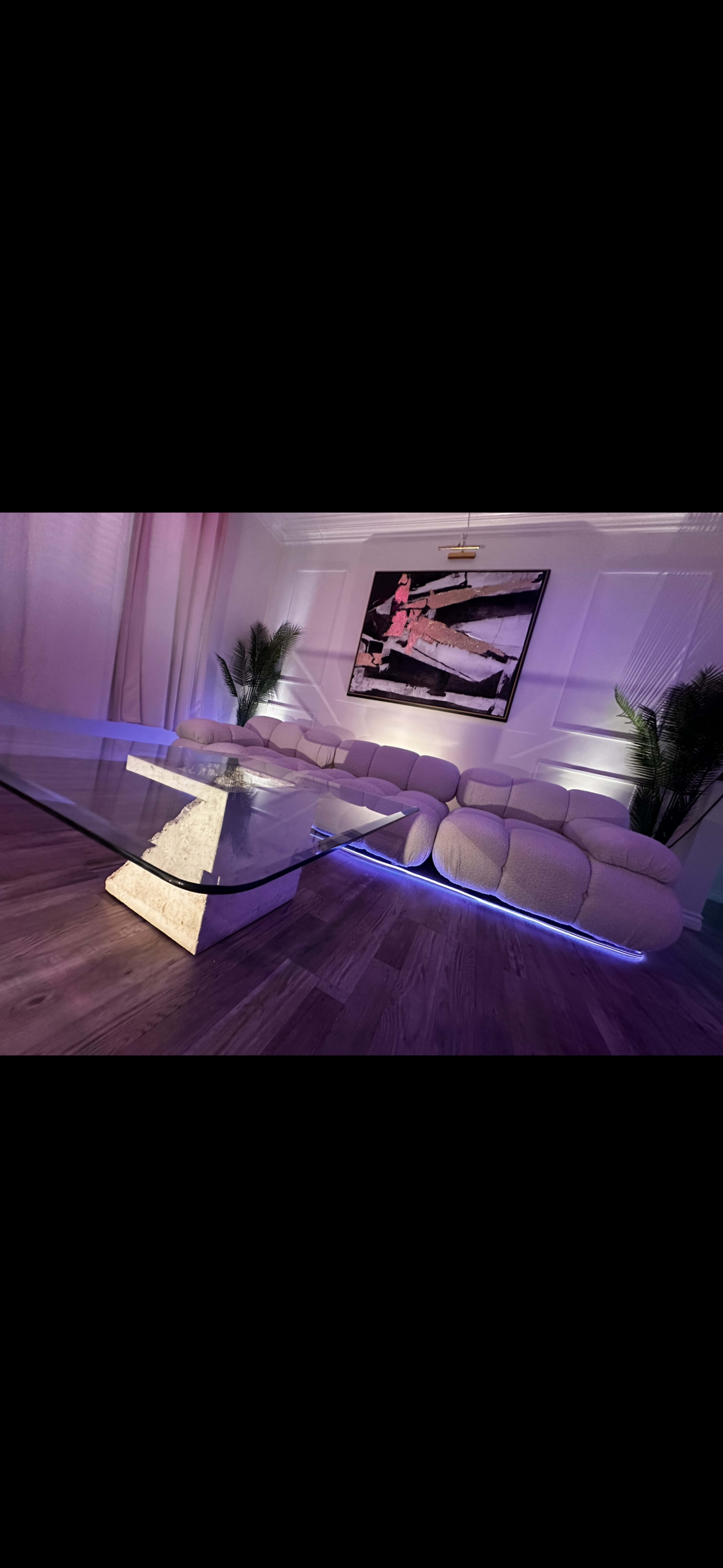 A modern living room features a light-colored sectional sofa, a glass coffee table with illuminated edges, and decorative plants against a backdrop of a large artwork.