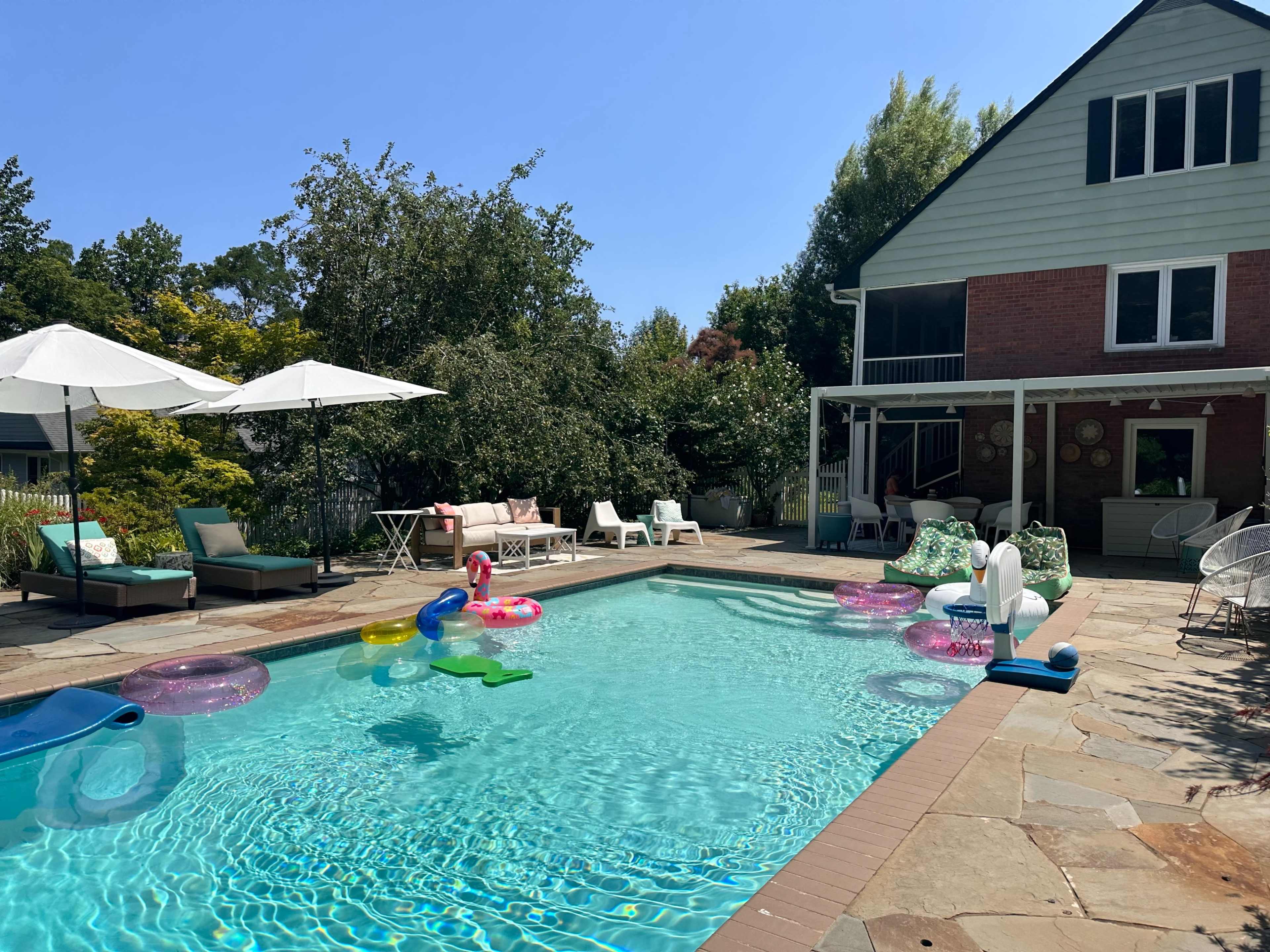 The image shows a backyard pool surrounded by lounge chairs, umbrellas, and colorful inflatable pool toys, with a house visible in the background.