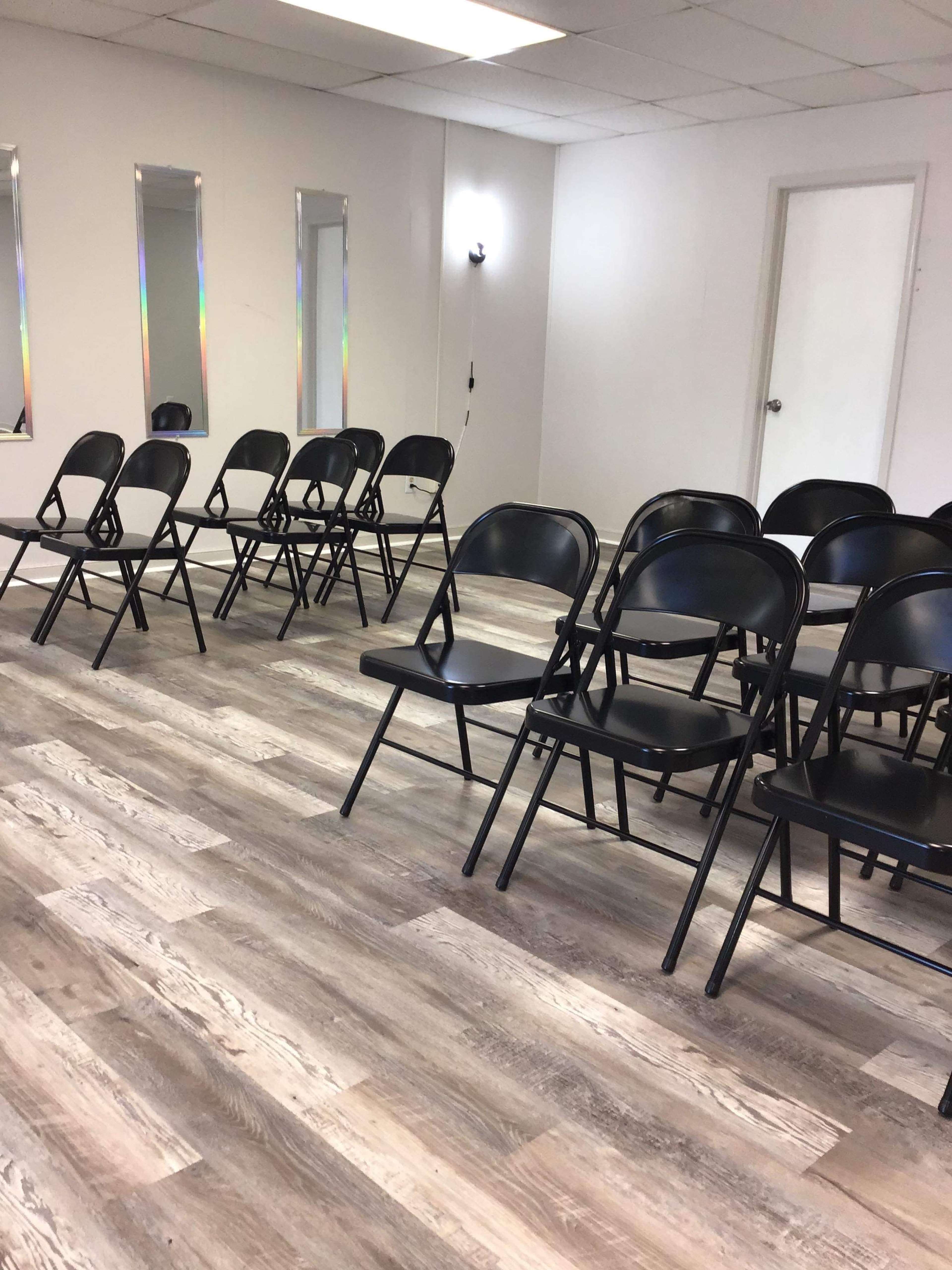 A room with several rows of black folding chairs arranged on a wooden floor, with a few narrow windows and a door visible in the background.