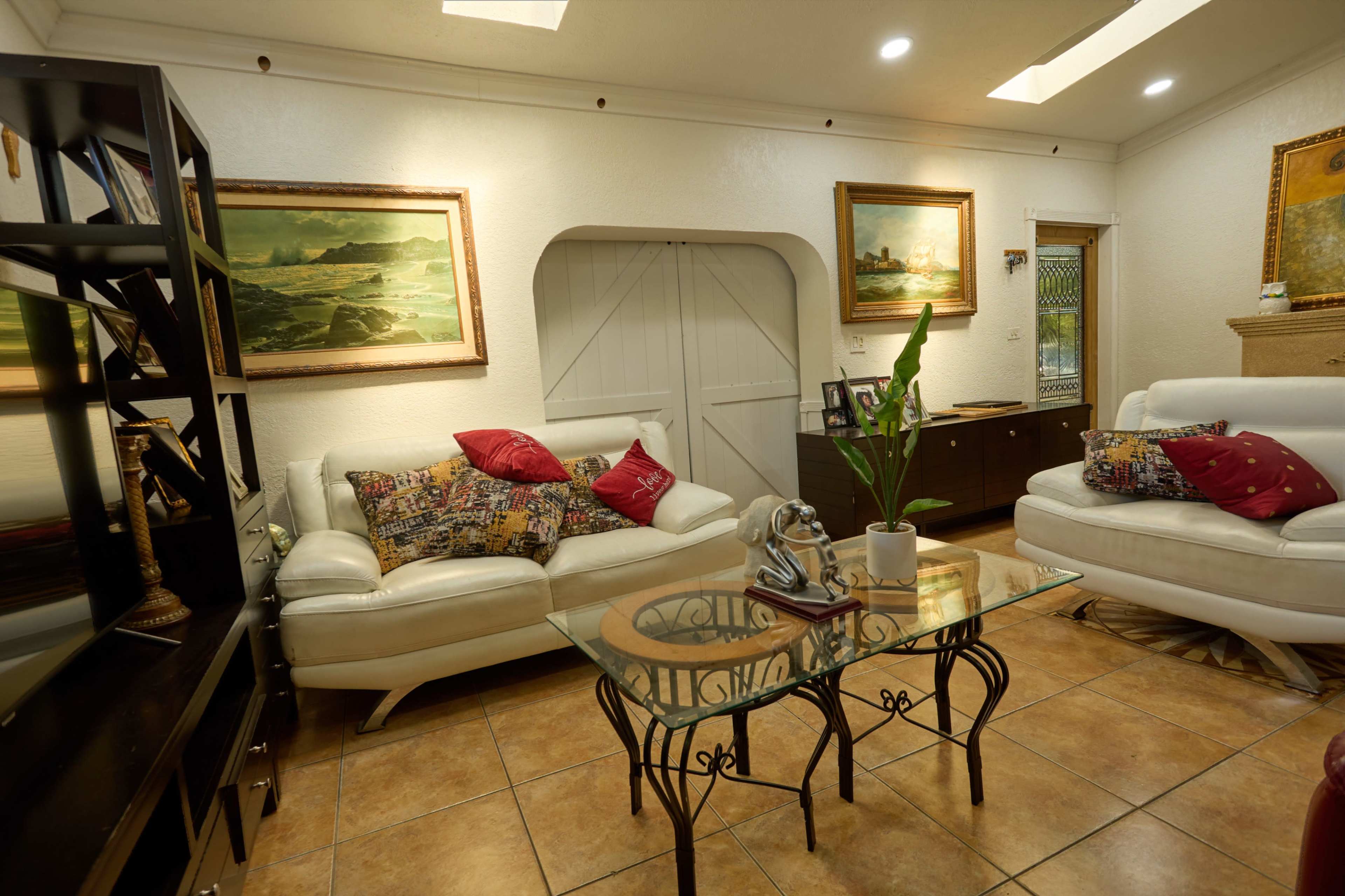 The image shows a well-furnished living room with white sofas, a glass coffee table, and paintings on the walls.
