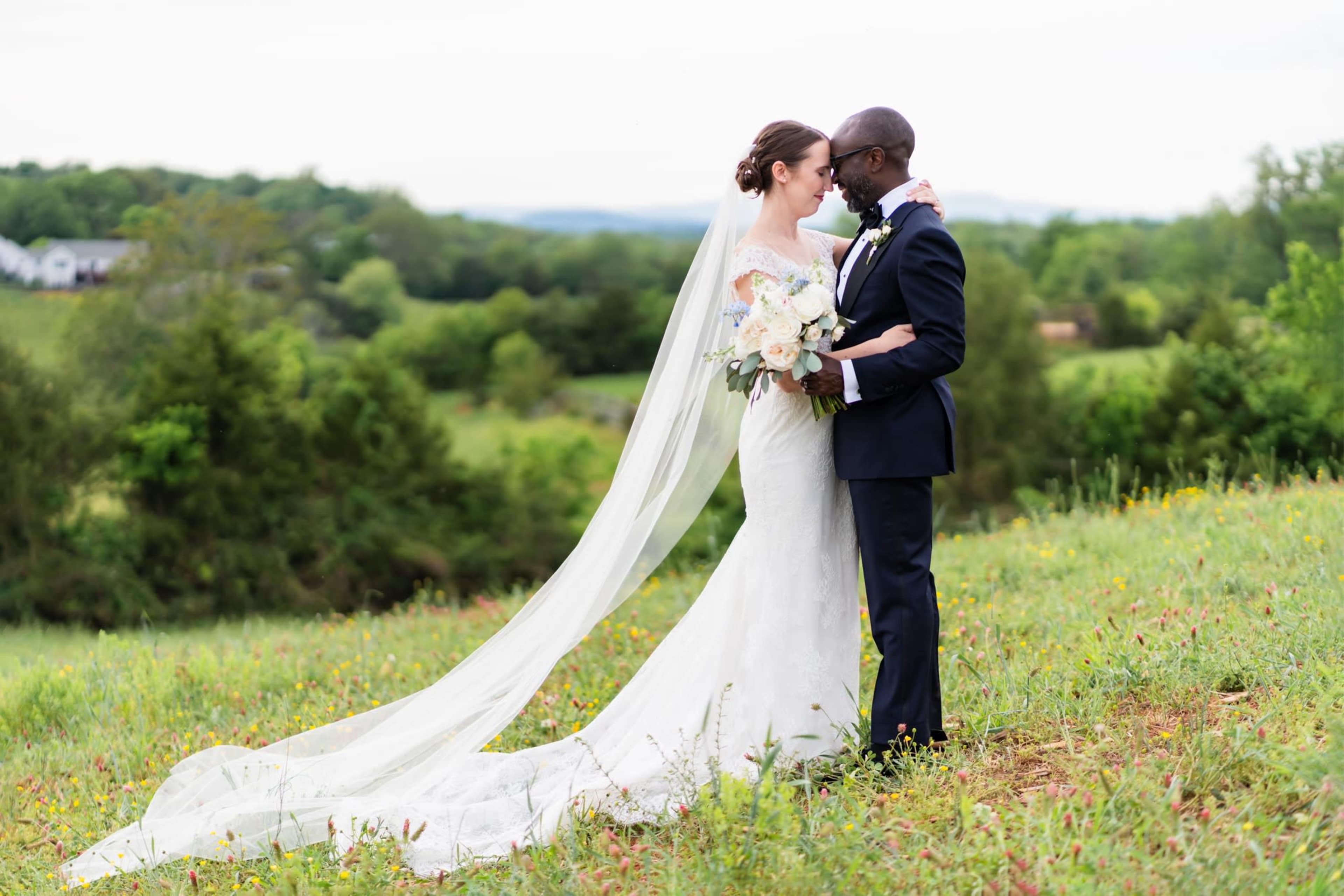A couple dressed in wedding attire stands in a grassy field, embracing each other with a scenic landscape in the background.