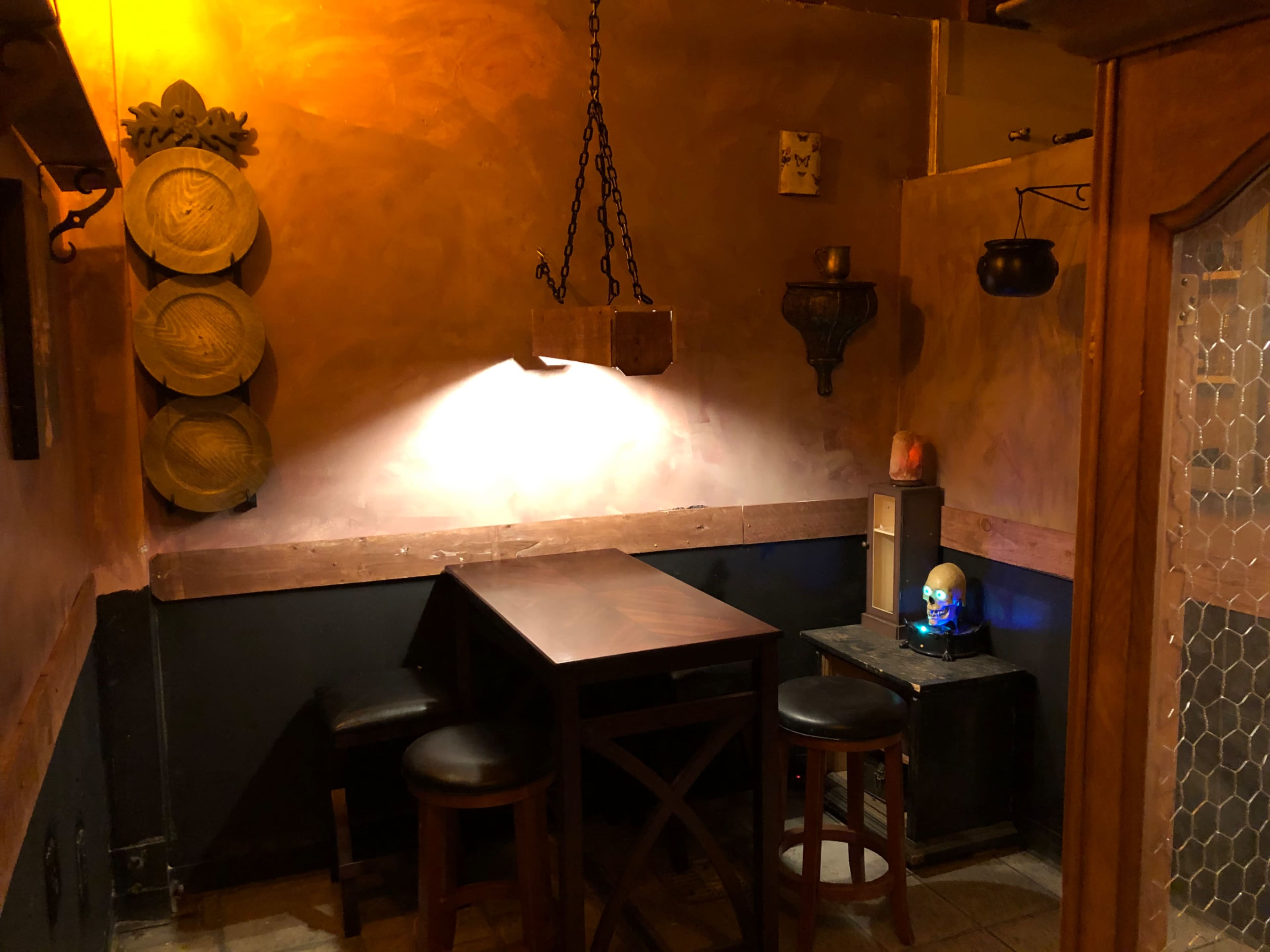The image shows a dimly lit corner of a bar or tavern with a wooden table and stools against a textured wall adorned with decorative items.