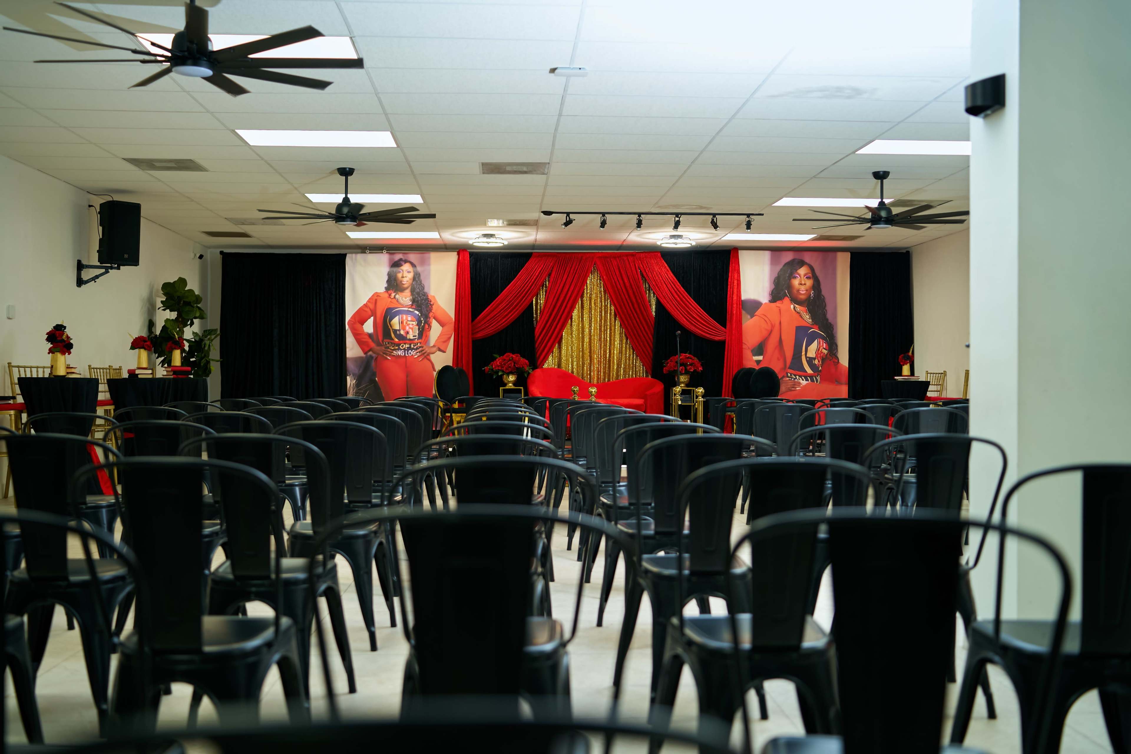The image shows a room set up for an event, featuring rows of black chairs facing a stage adorned with red and gold decorations and large portraits.