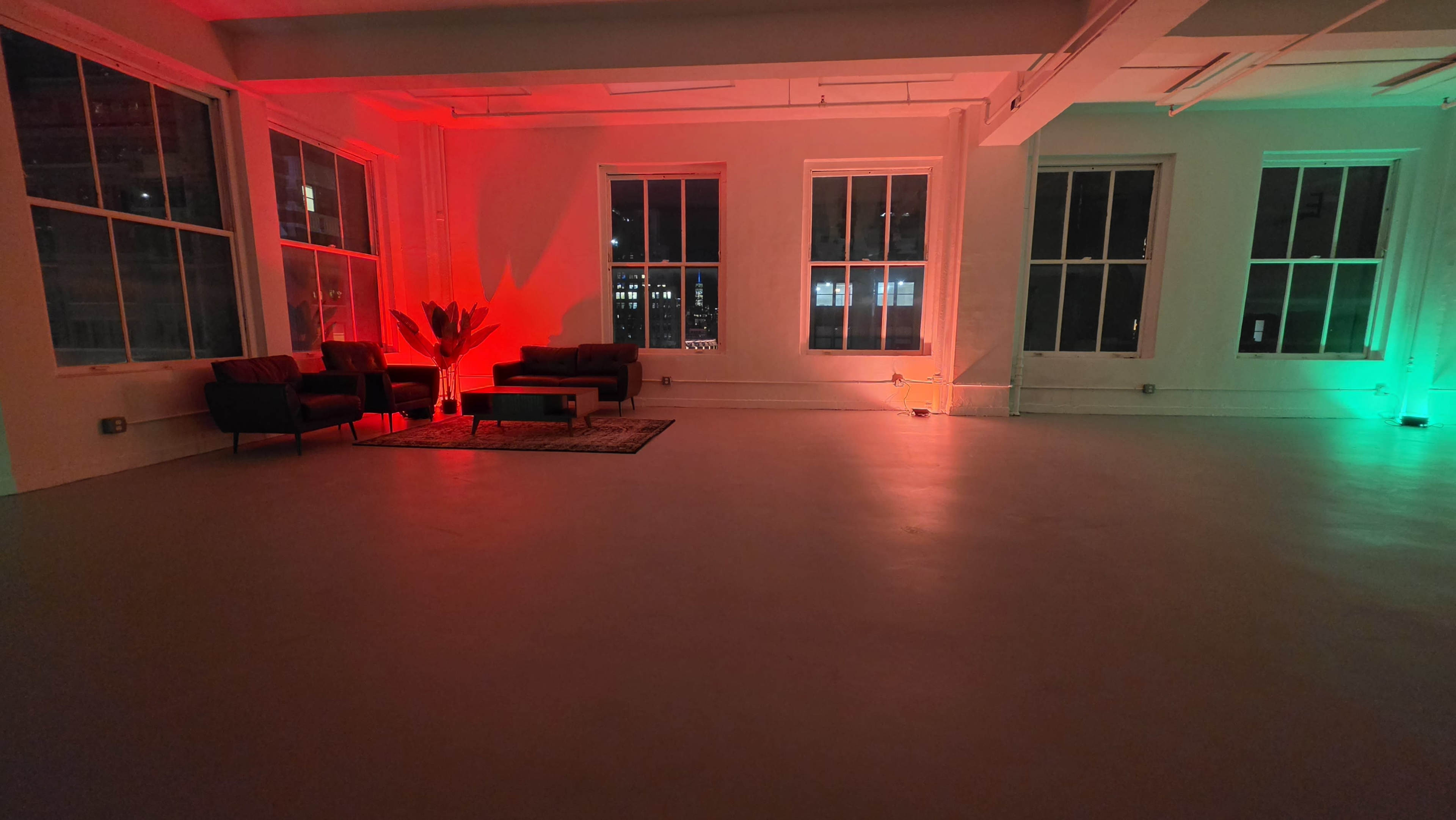 The image shows an empty room with red and green lighting, featuring a couple of dark couches and large windows.