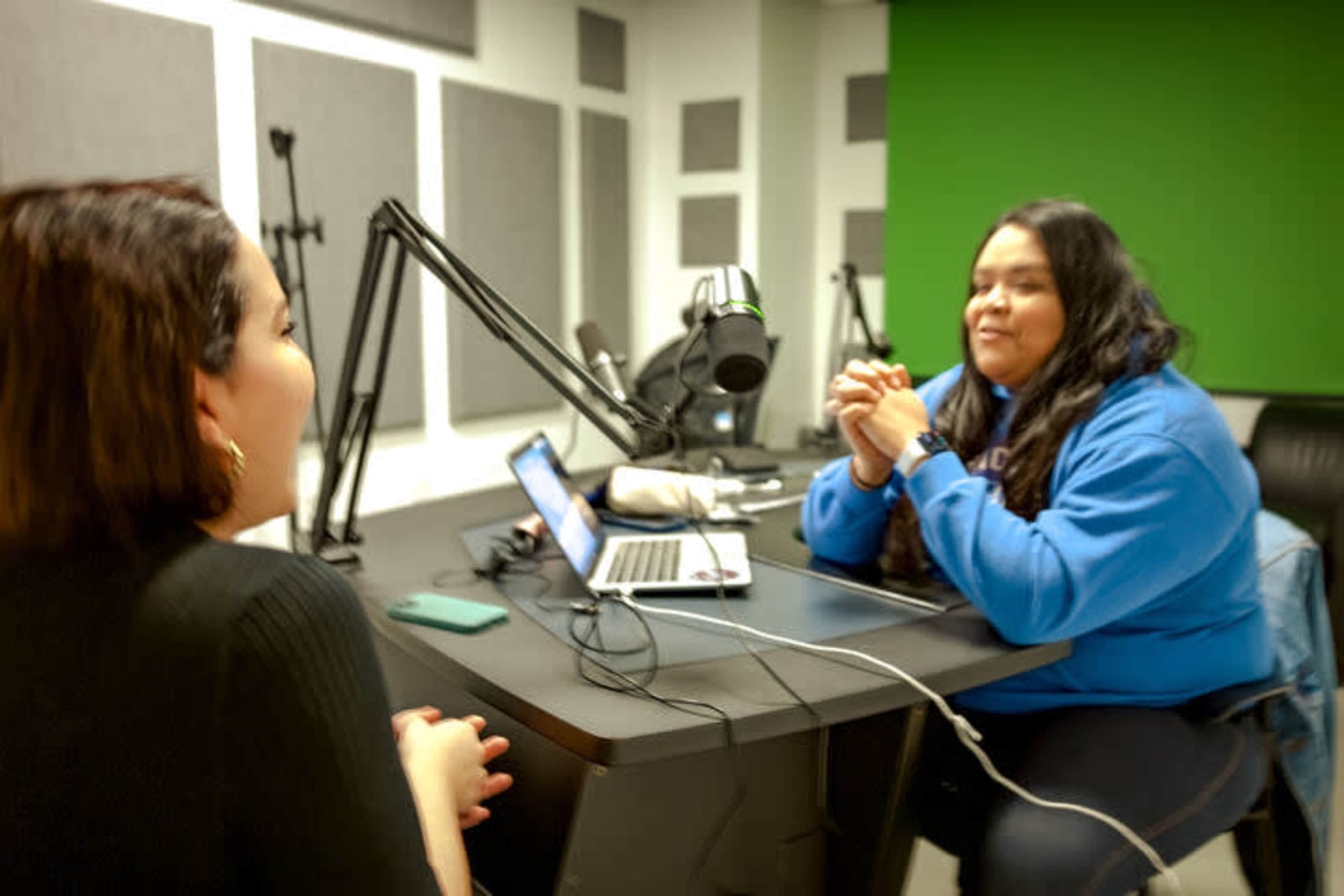 Two women are seated at a table in a recording studio equipped with a microphone and a laptop.