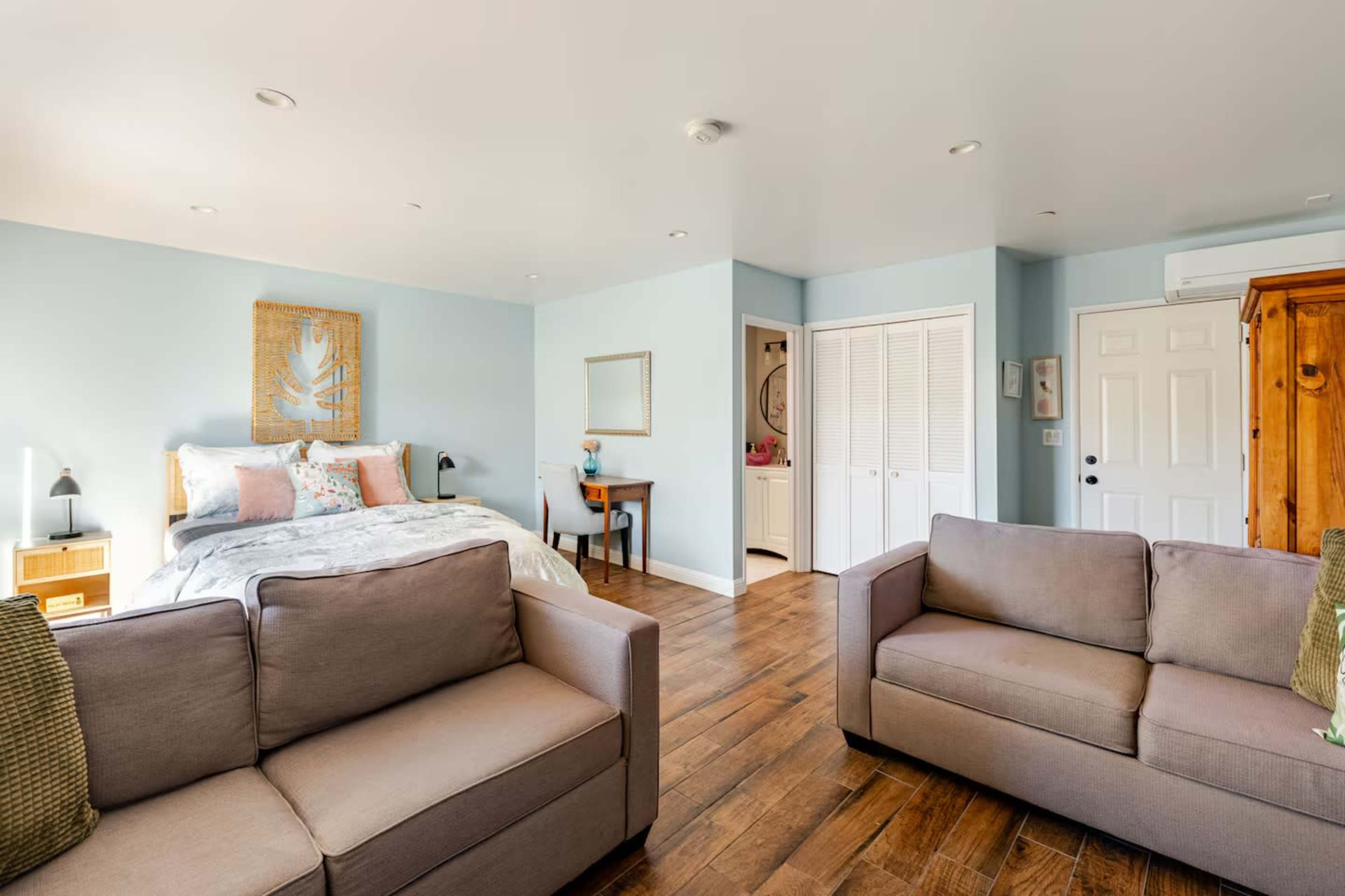 The image shows a bright, airy room featuring a bed with decorative pillows, a small table with a mirror, and two upholstered couches arranged in the living area.