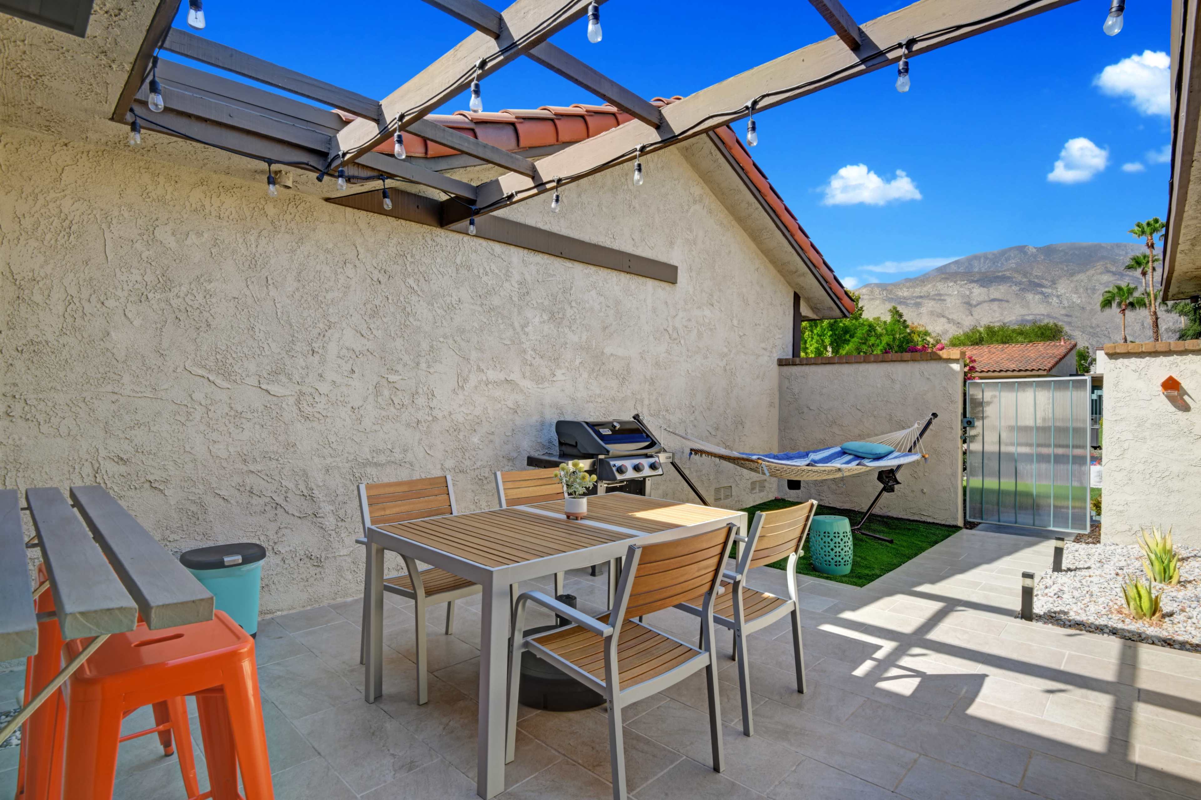 A patio area features a table and chairs under a covered terrace, with a grill, a hammock, and a view of mountains in the background.
