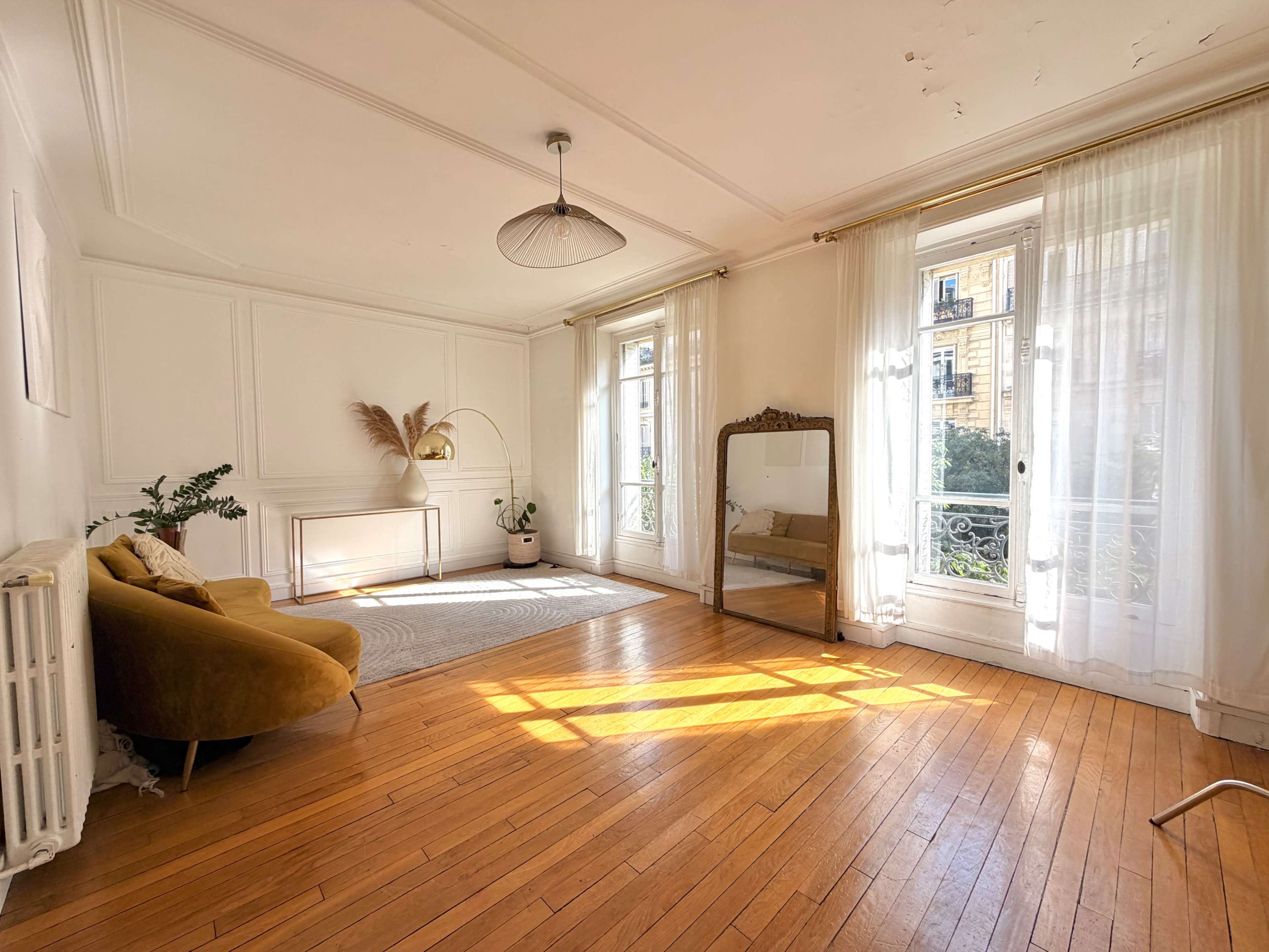 The image shows a bright room with wooden flooring, large windows with sheer curtains, a yellow sofa, a floor mirror, and a decorative plant.