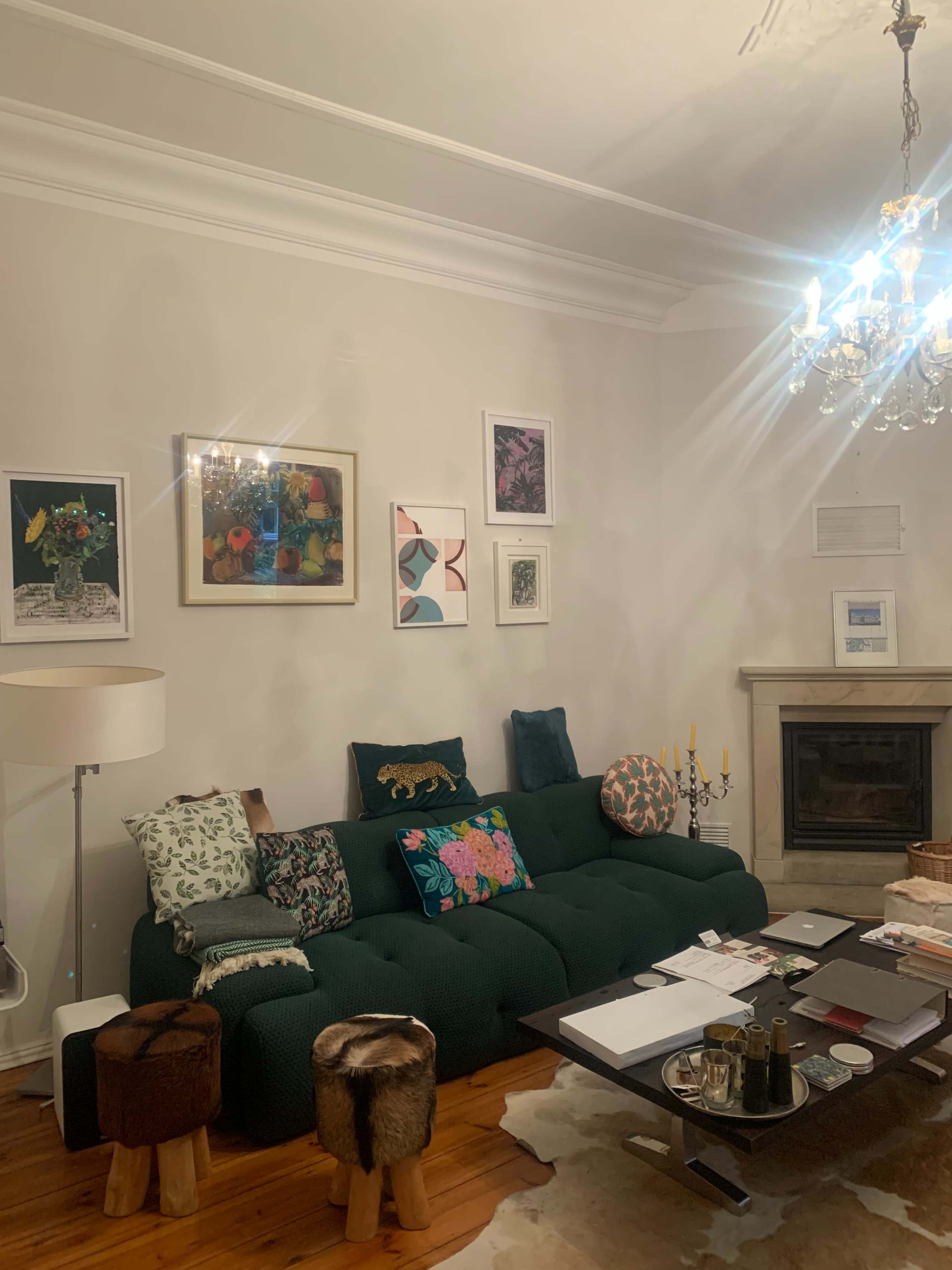 A cozy living room features a green sofa adorned with colorful cushions, a coffee table cluttered with books and decorative items, and various artworks displayed on the walls.