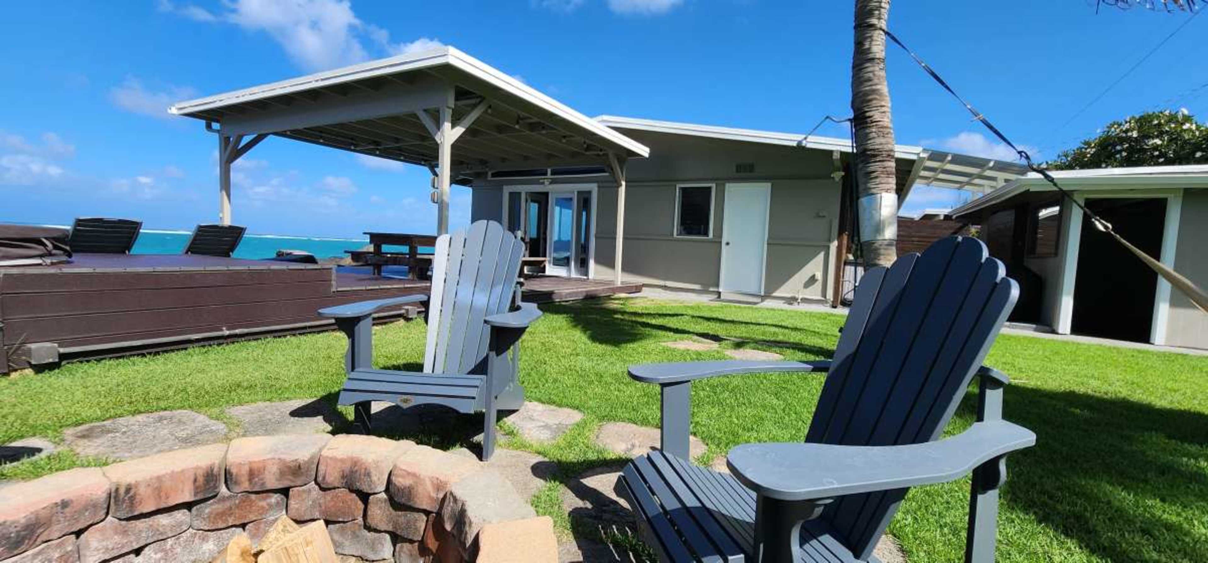 A seaside house features a grassy area with two gray Adirondack chairs facing a circular stone fire pit under a blue sky.