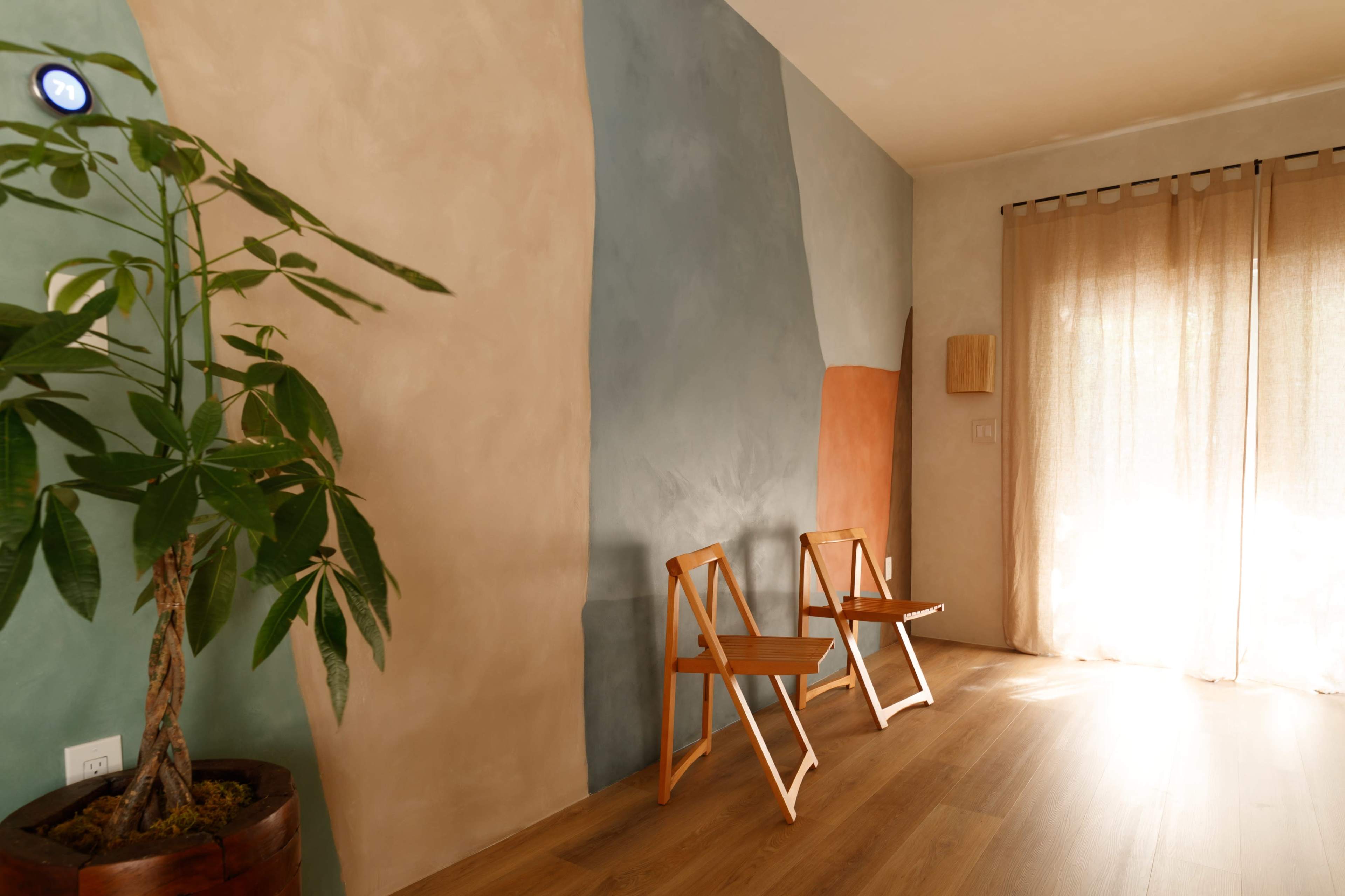 The image shows a room with two wooden folding chairs against a wall featuring three different colored sections, a potted plant in the foreground, and light filtering through sheer curtains.