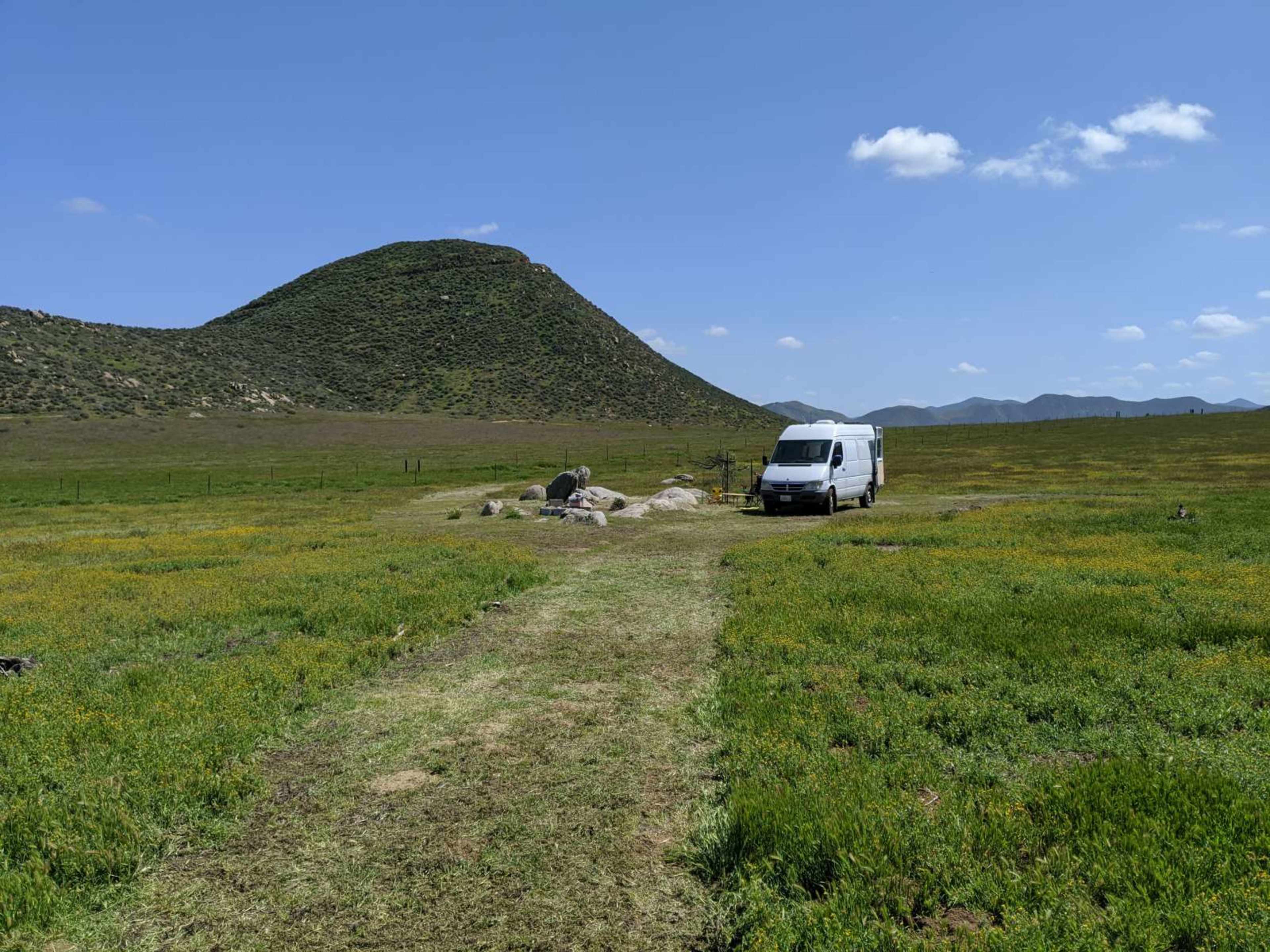 A white van is parked on a dirt path surrounded by green fields and a large hill in the background under a blue sky.
