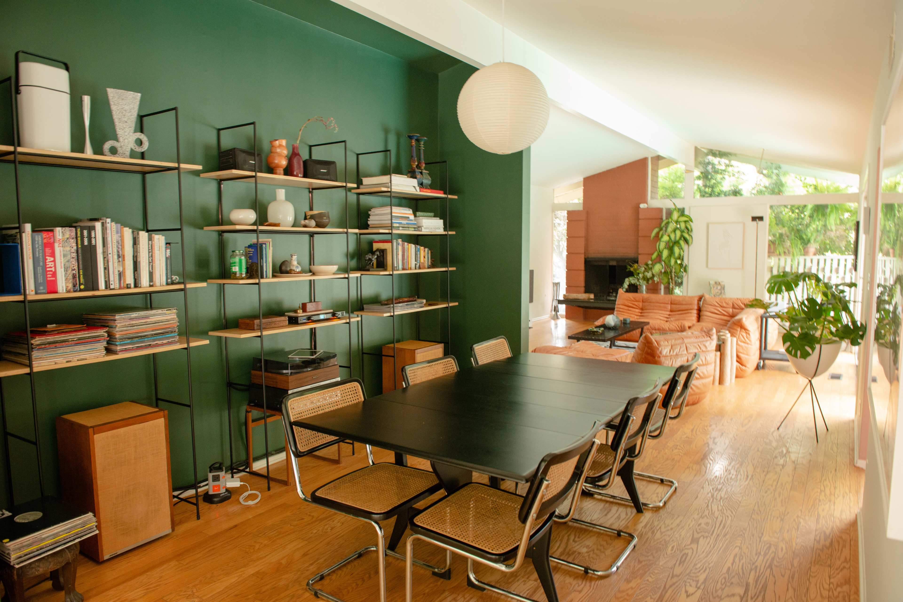 A modern dining area with a black table and chairs, a green wall featuring shelves filled with books and decorative items, and a cozy lounge area visible in the background.