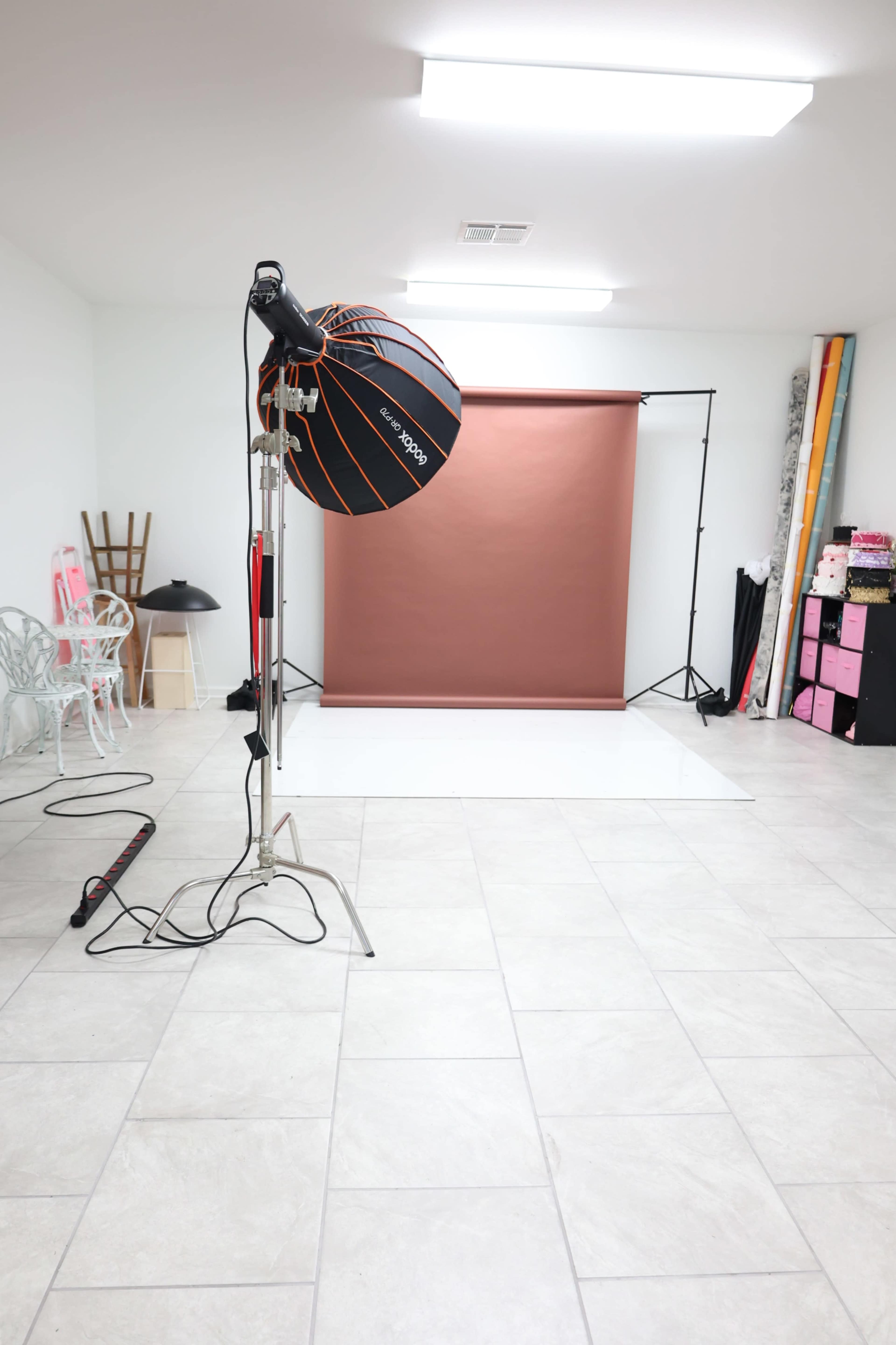 The image shows a photography studio with a backdrop setup, lighting equipment, and various props arranged in a well-lit room with tile flooring.