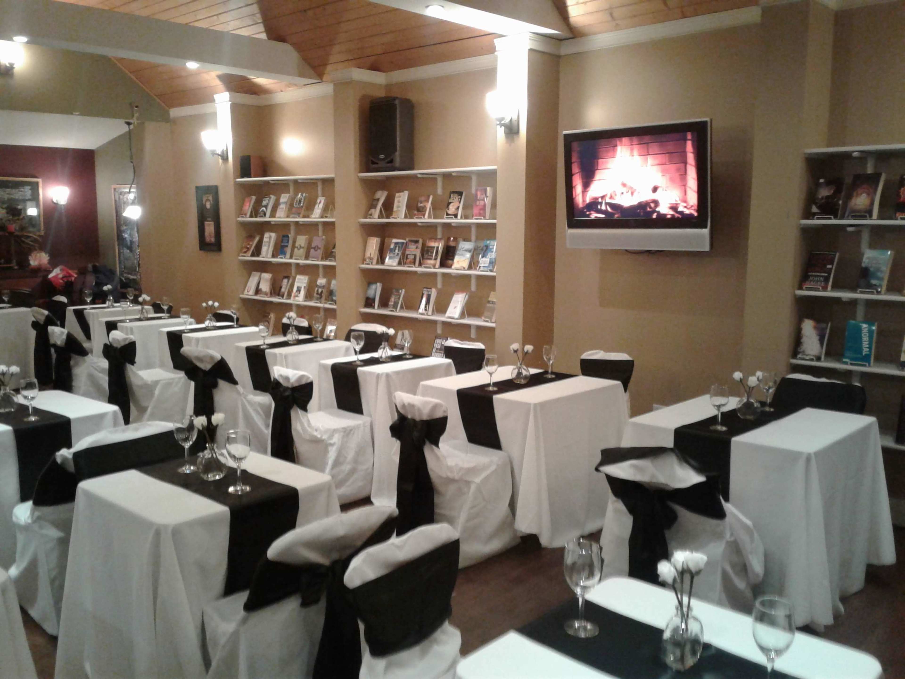 A dining area with white tablecloths and black bows on chairs, featuring shelves of books and a television displaying a fireplace.