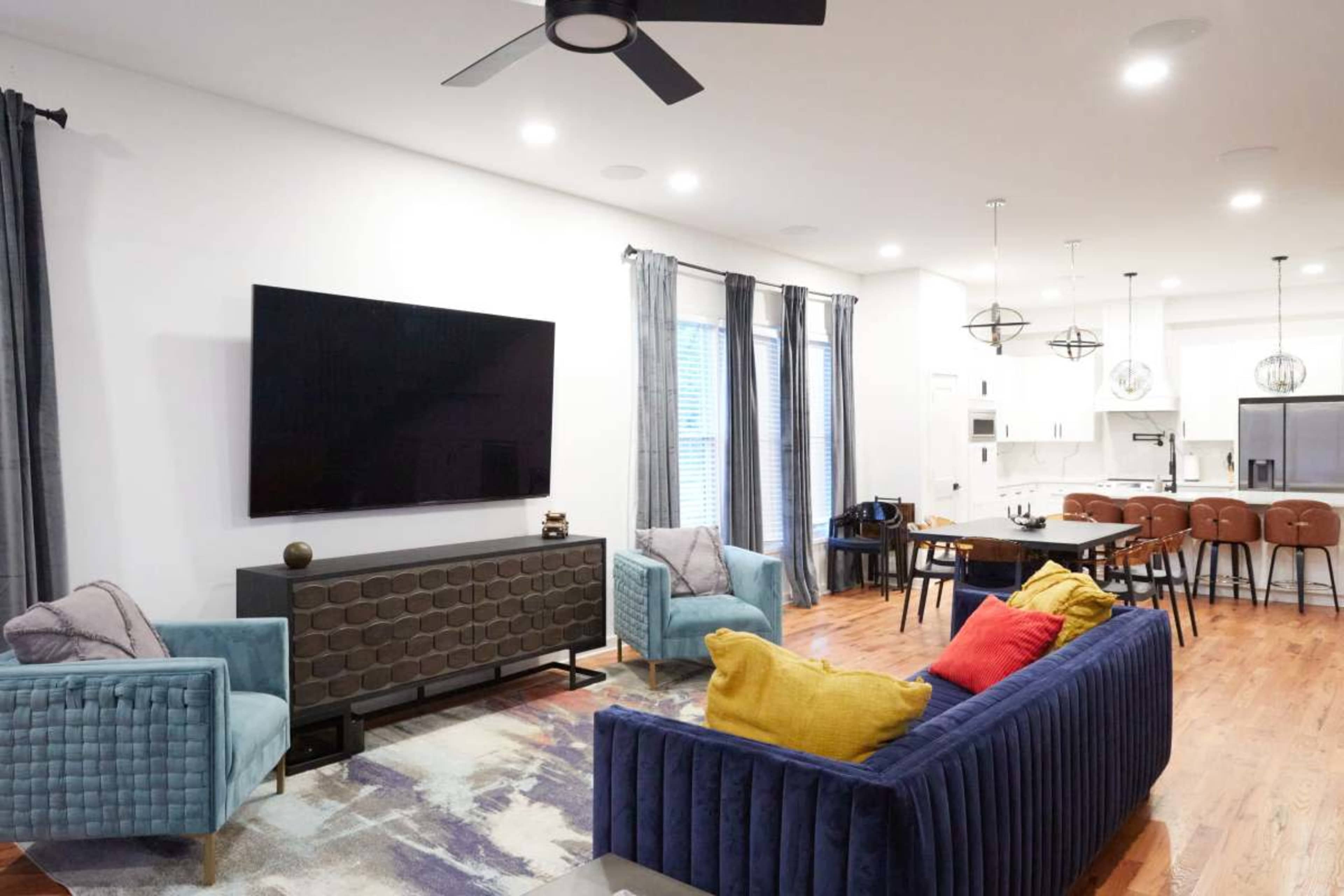 The image shows a modern living room with two upholstered sofas, a large wall-mounted television, a dining area with a table and chairs, and wood flooring.