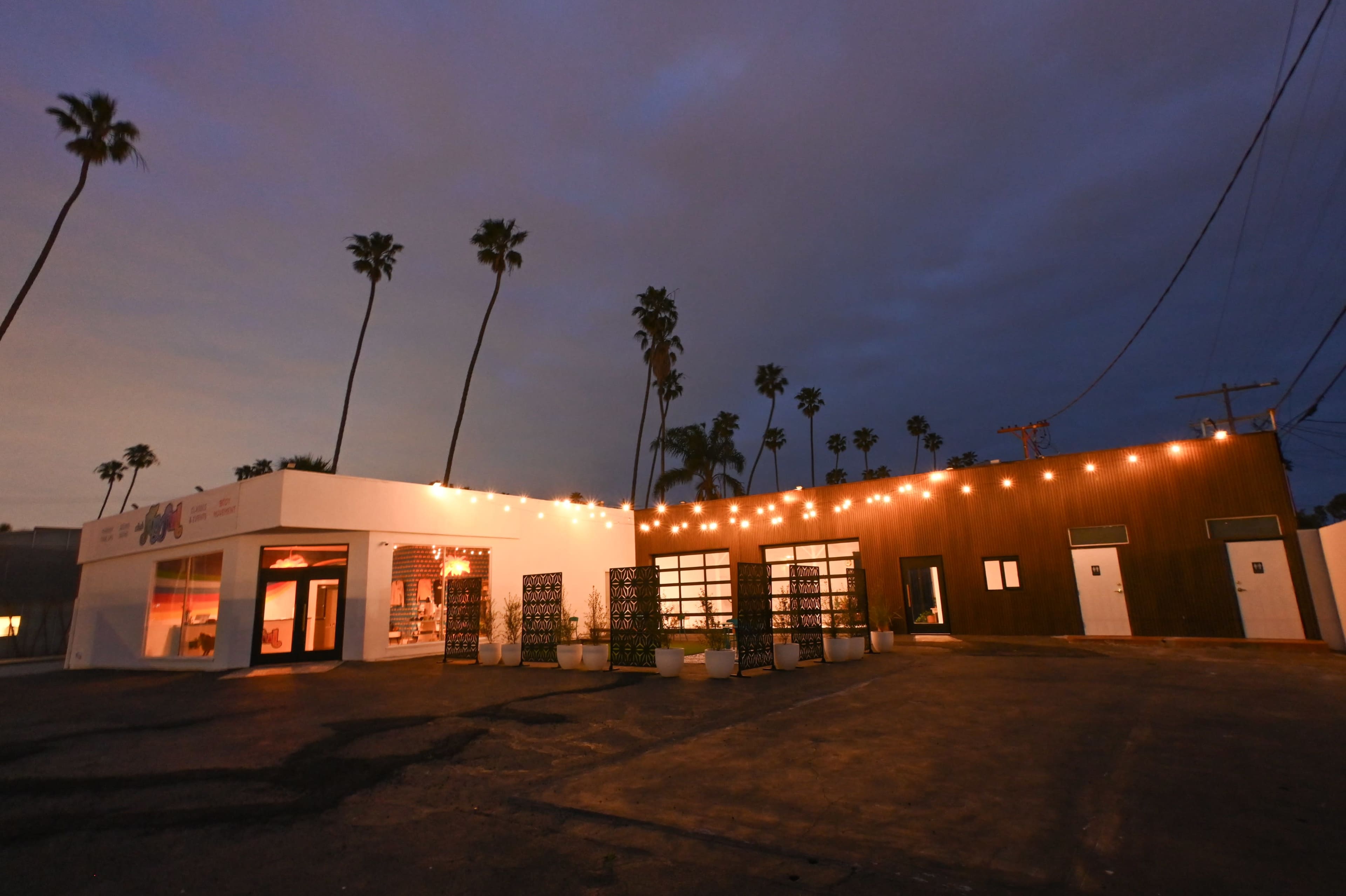 A modern building with string lights along the roof, surrounded by palm trees under a twilight sky.