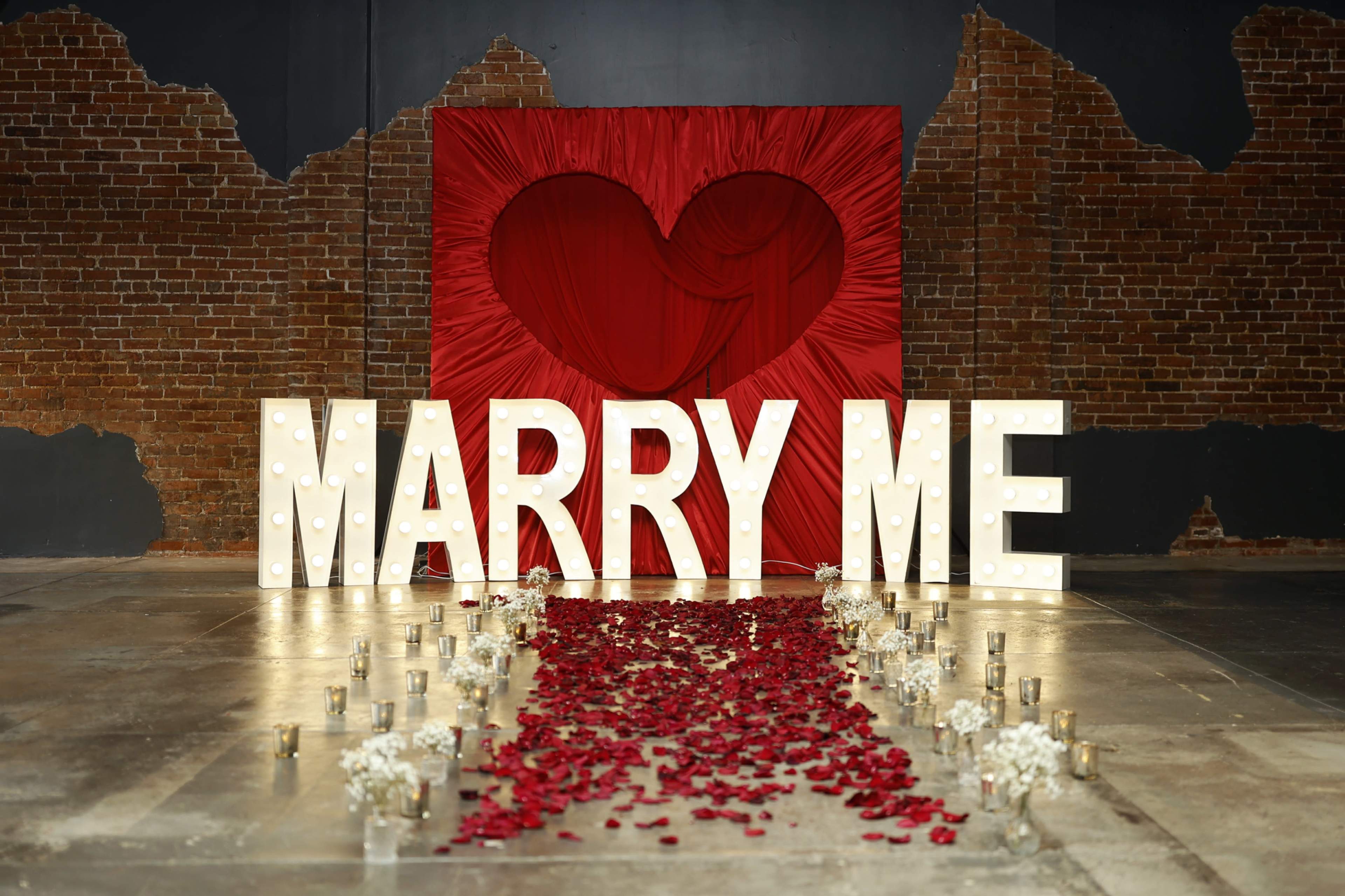 The scene features large illuminated letters spelling "MARRY ME" in front of a red heart backdrop, surrounded by a path of rose petals and small candles.