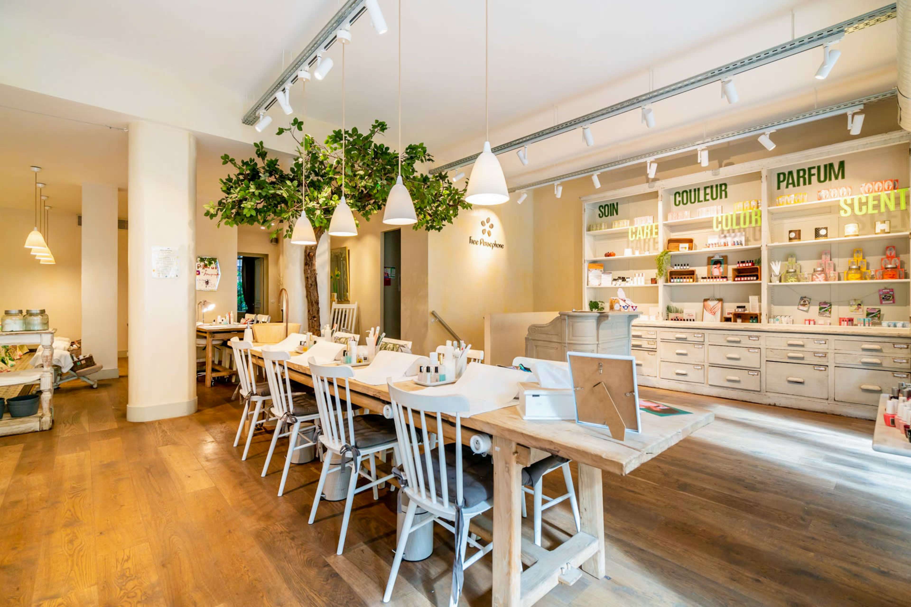 A spacious and well-lit interior features a wooden communal table surrounded by white chairs, with shelves displaying various products in the background.