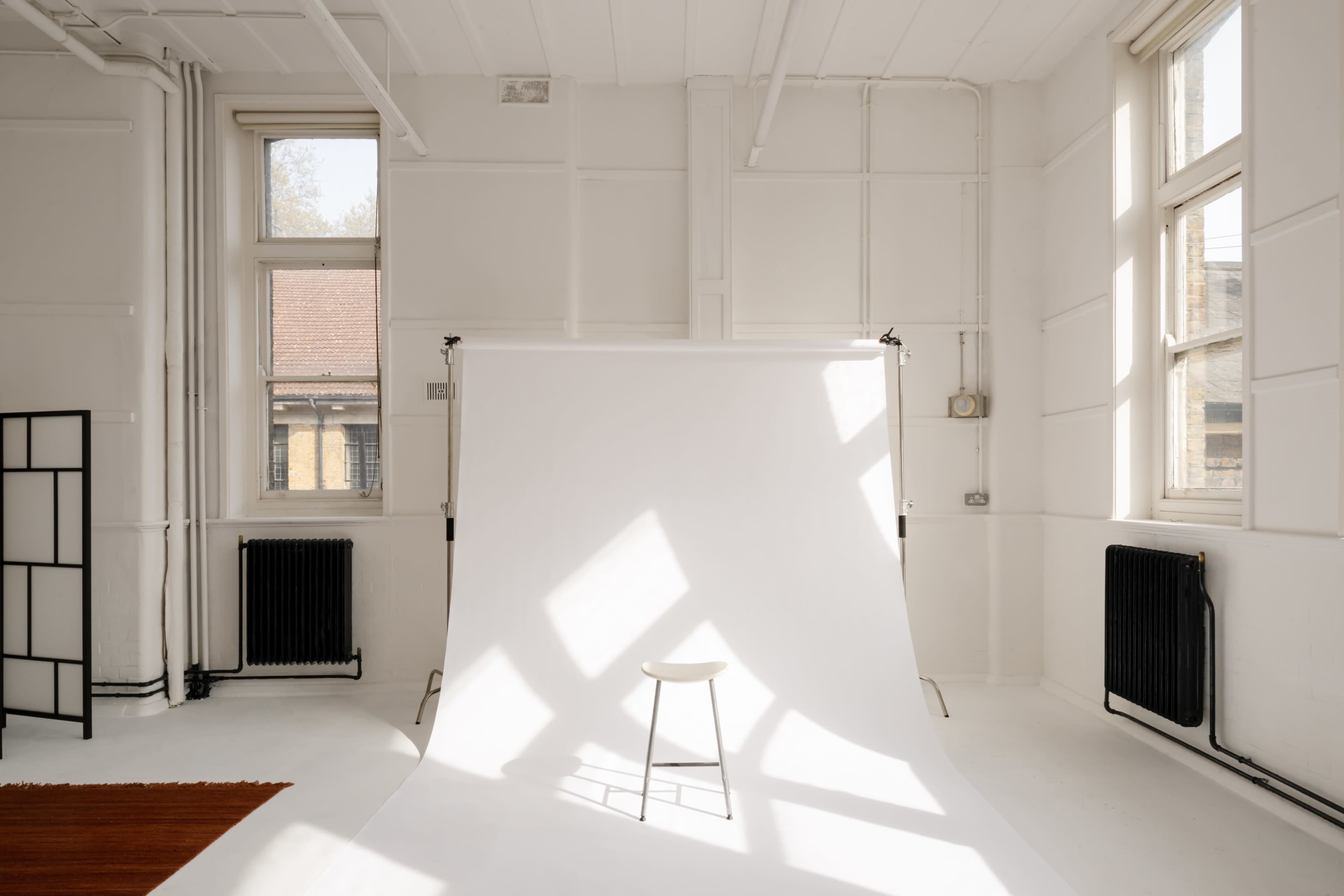 A minimalist photography studio features a white backdrop and a single wooden stool under natural light.