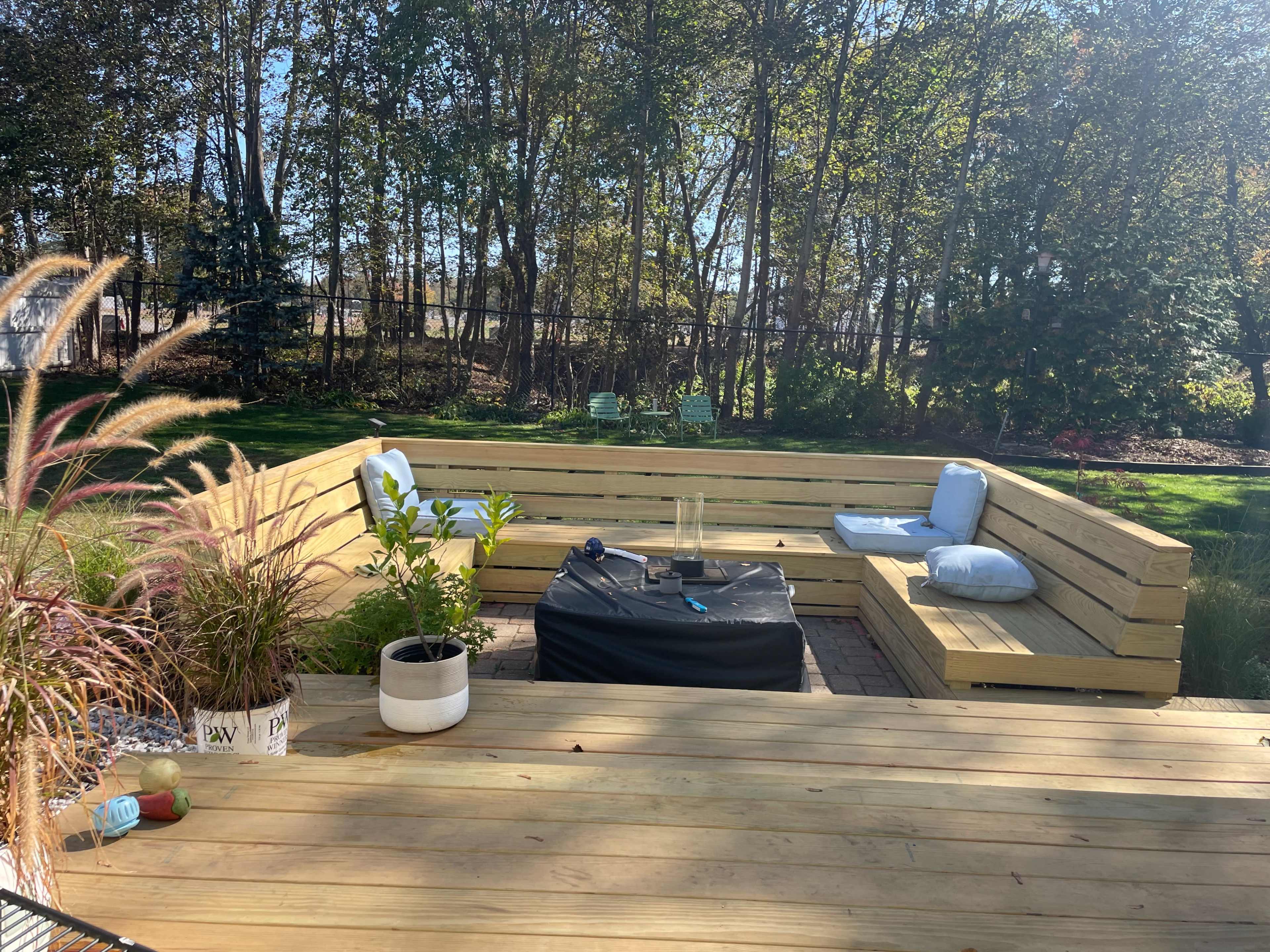The image shows a wooden patio with a sectional seating area, a coffee table, and potted plants, all surrounded by trees in a sunlit outdoor environment.