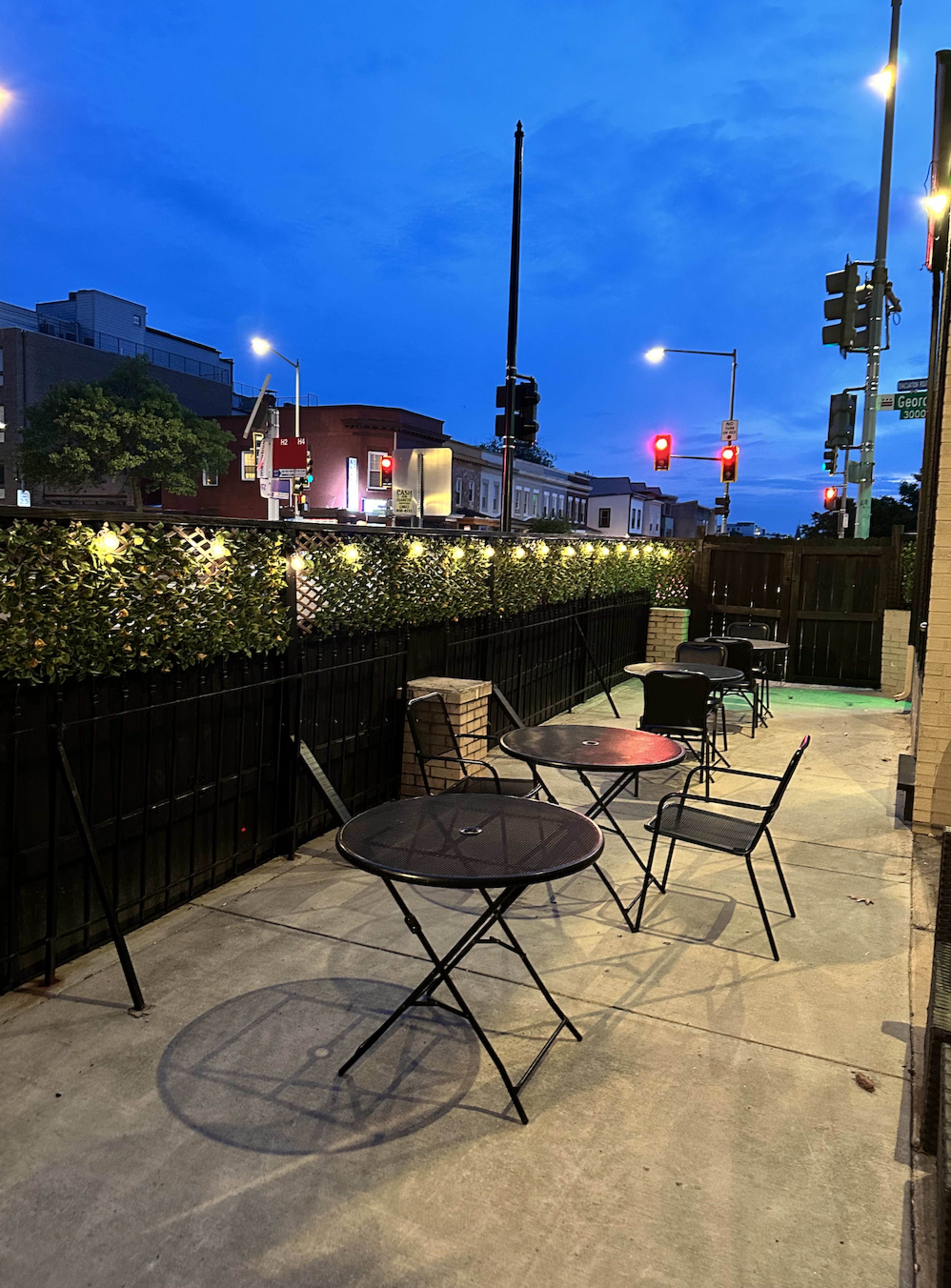 The image shows an outdoor seating area with several black metal tables and chairs, bordered by a low hedge, under a twilight sky with traffic lights visible in the background.