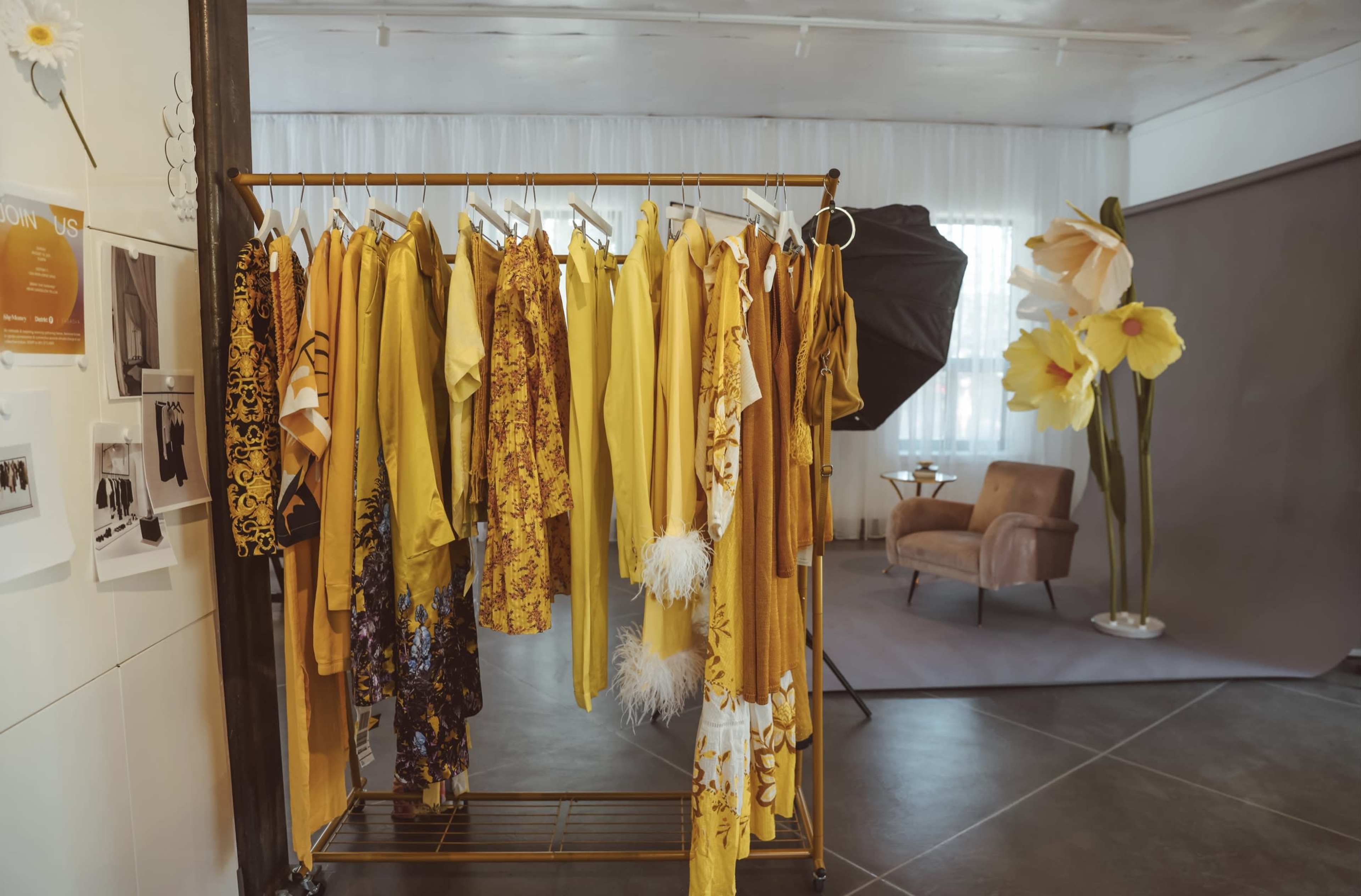 A clothing rack filled with various yellow garments stands in a well-lit studio space, alongside a beige armchair and large flower decorations.