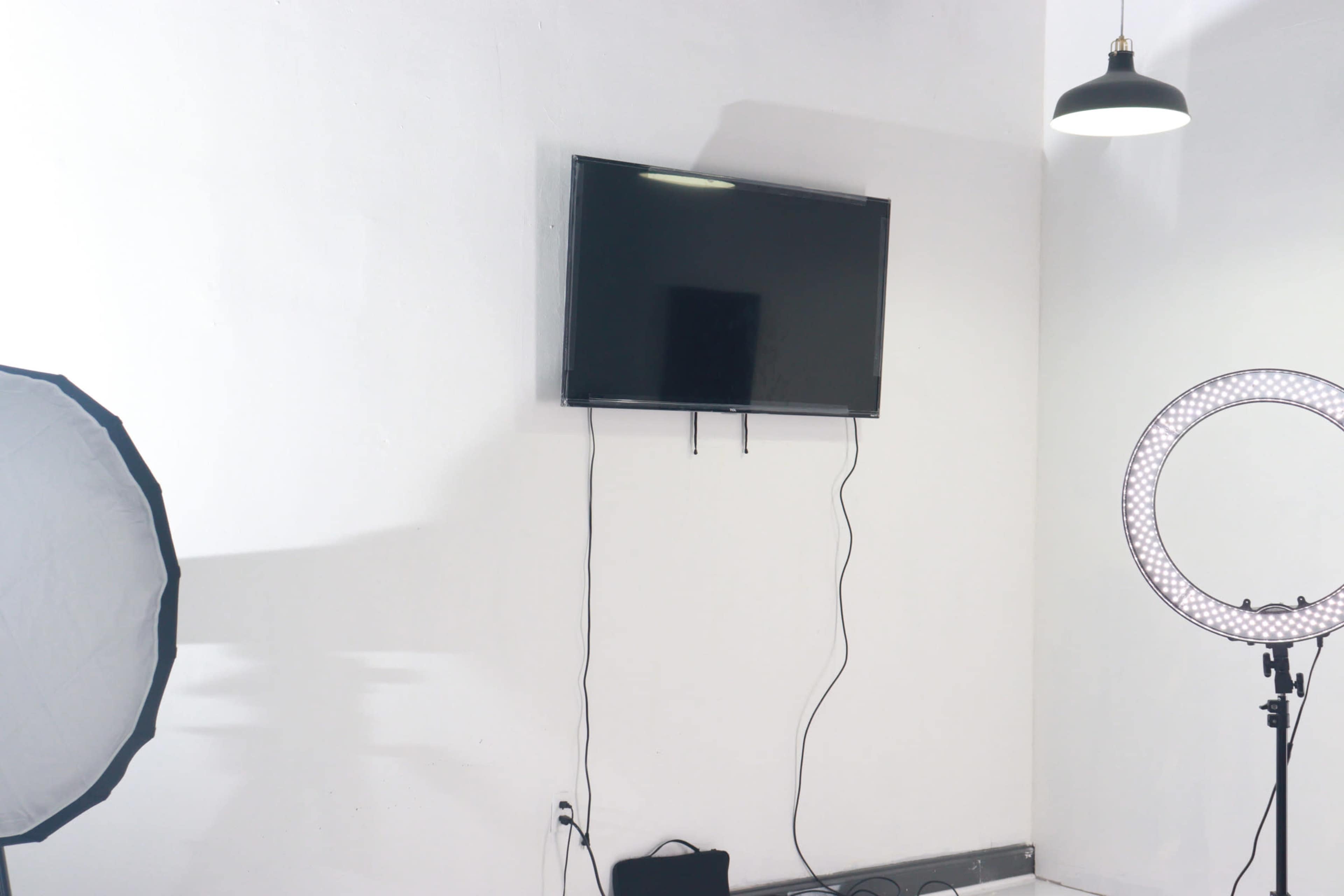 The image shows a television mounted on a white wall, alongside a ring light and softbox lighting setup in a studio-like environment.
