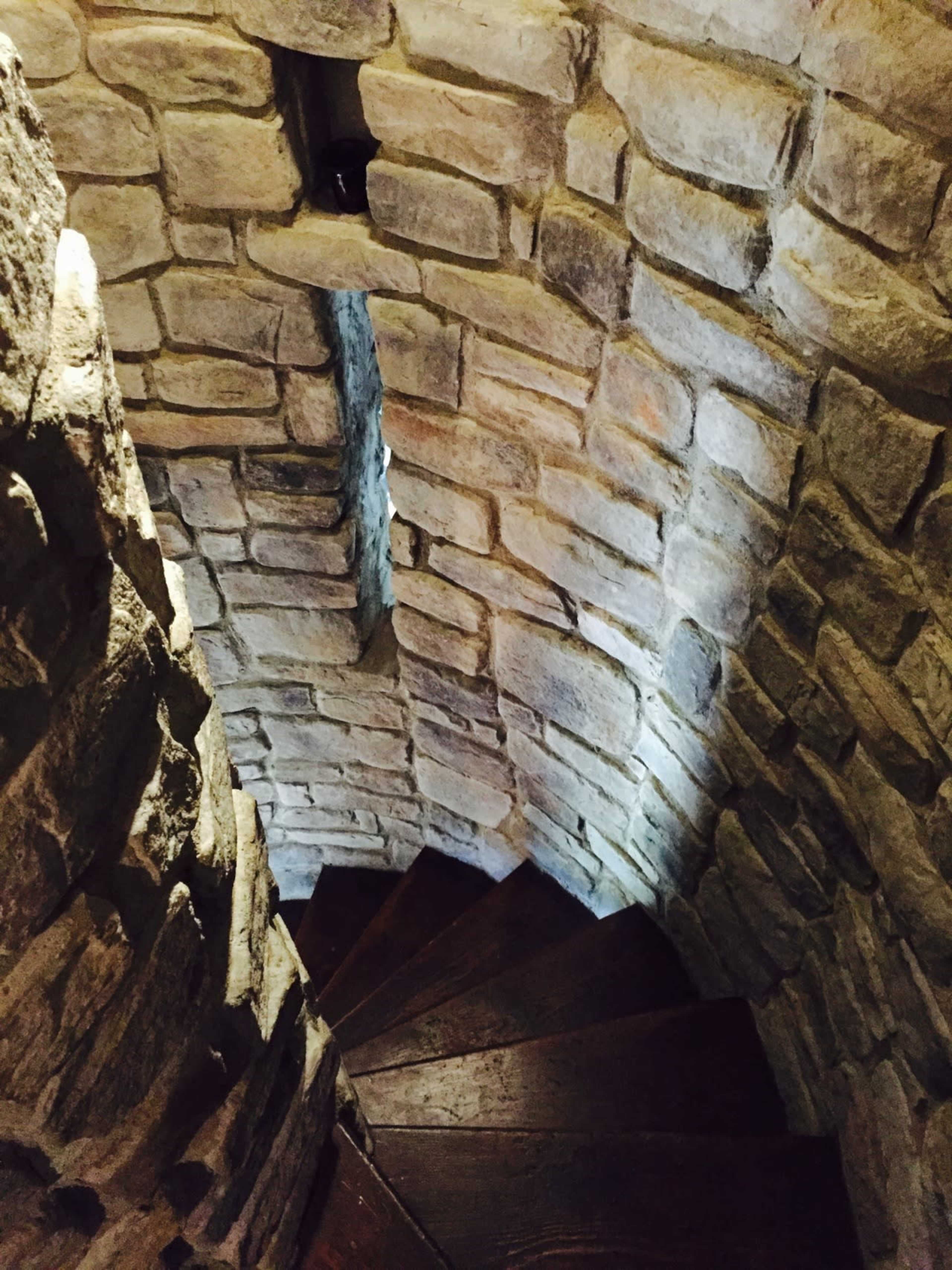 The image shows a winding stone staircase with wooden steps, enclosed by rough, textured stone walls.