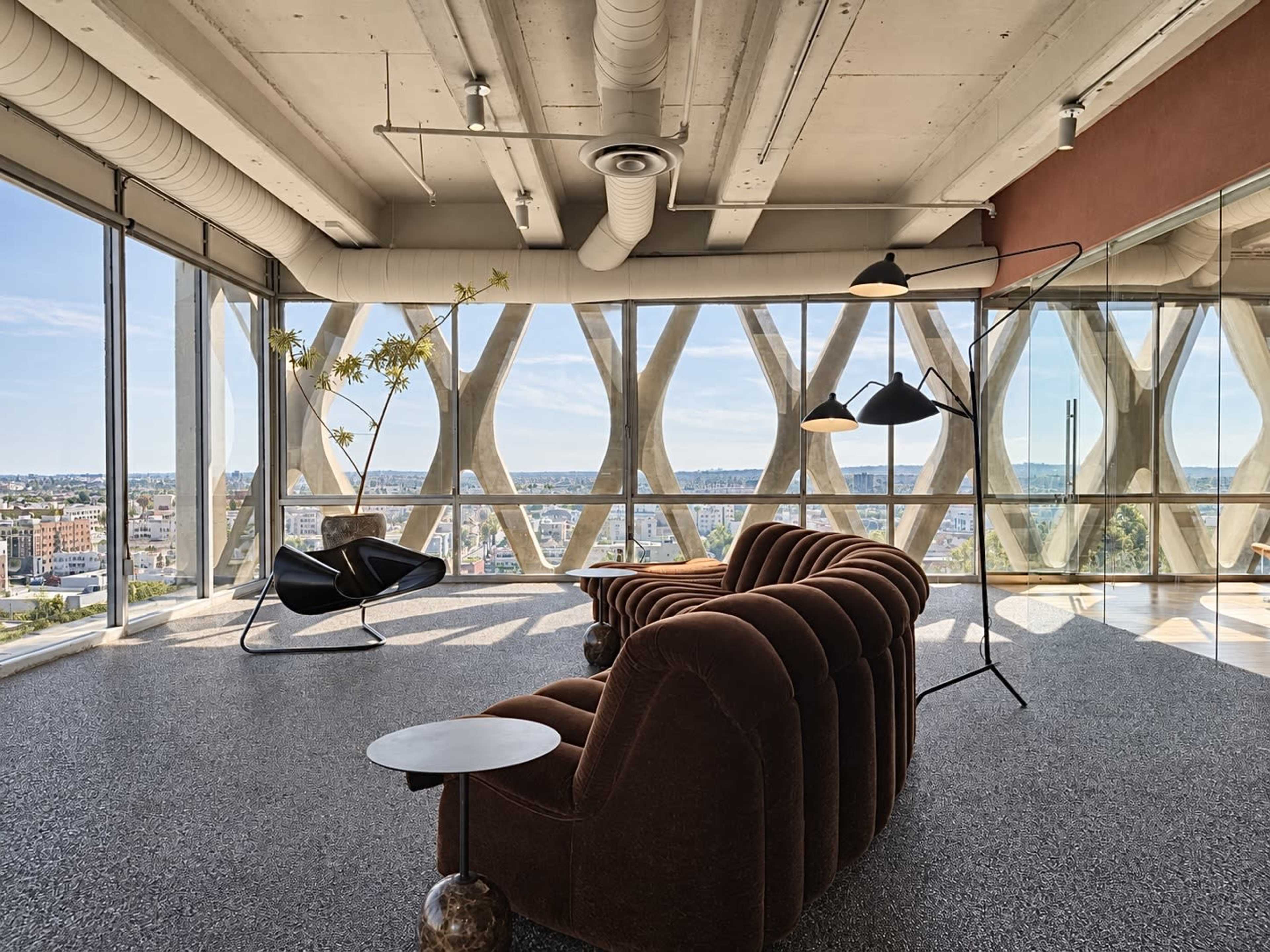 A modern interior space features a brown tufted sofa, a minimalist side table, and large windows offering a panoramic view of the city.