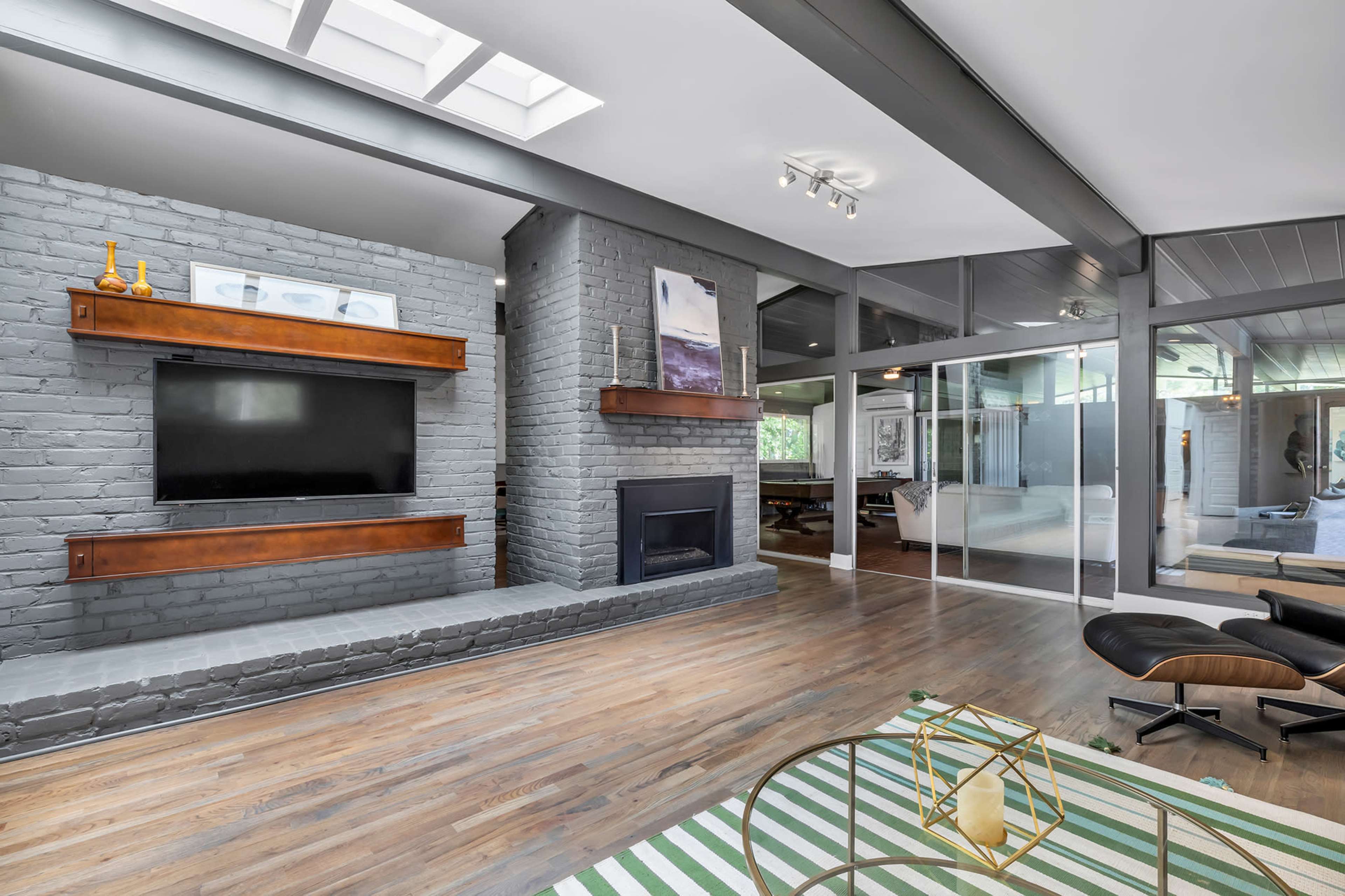 A modern living room with a gray brick fireplace, a mounted television, and large glass windows.