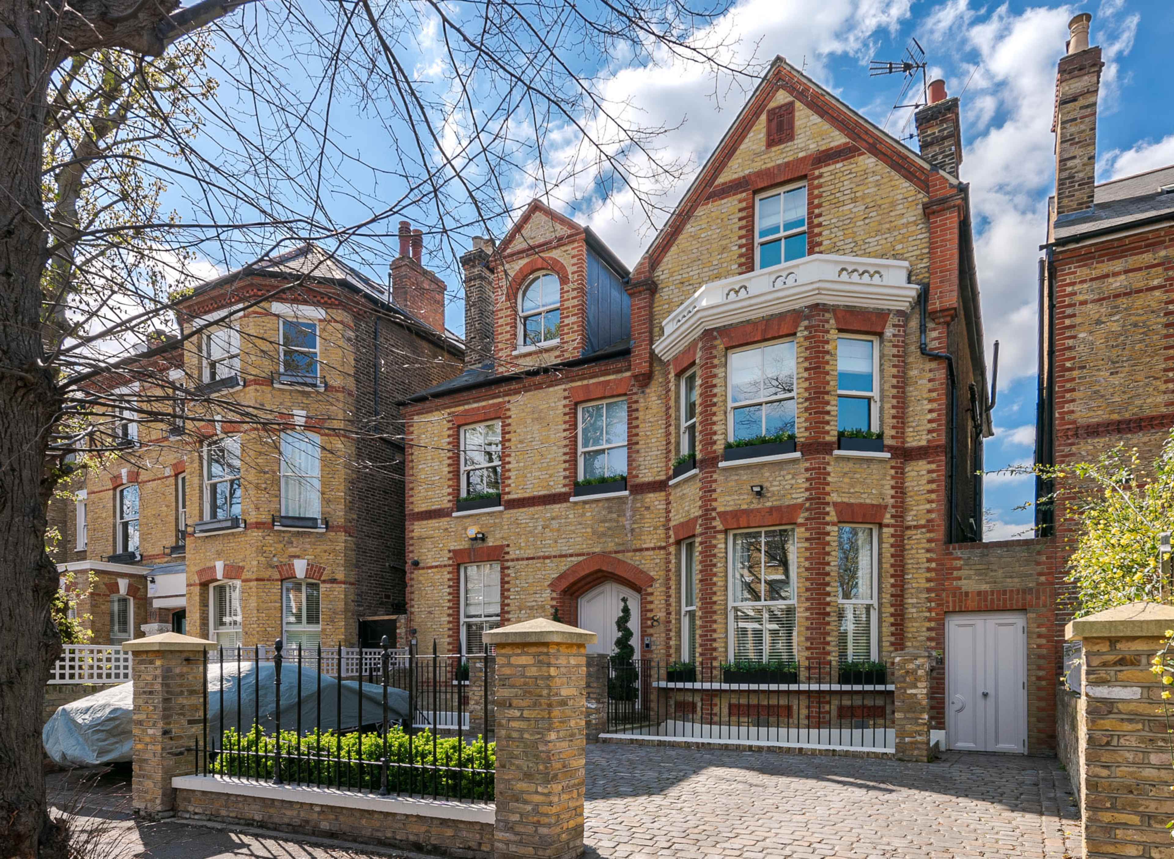 The image shows a brick house with a distinctive architectural style, flanked by similar buildings and surrounded by trees and paving stones.