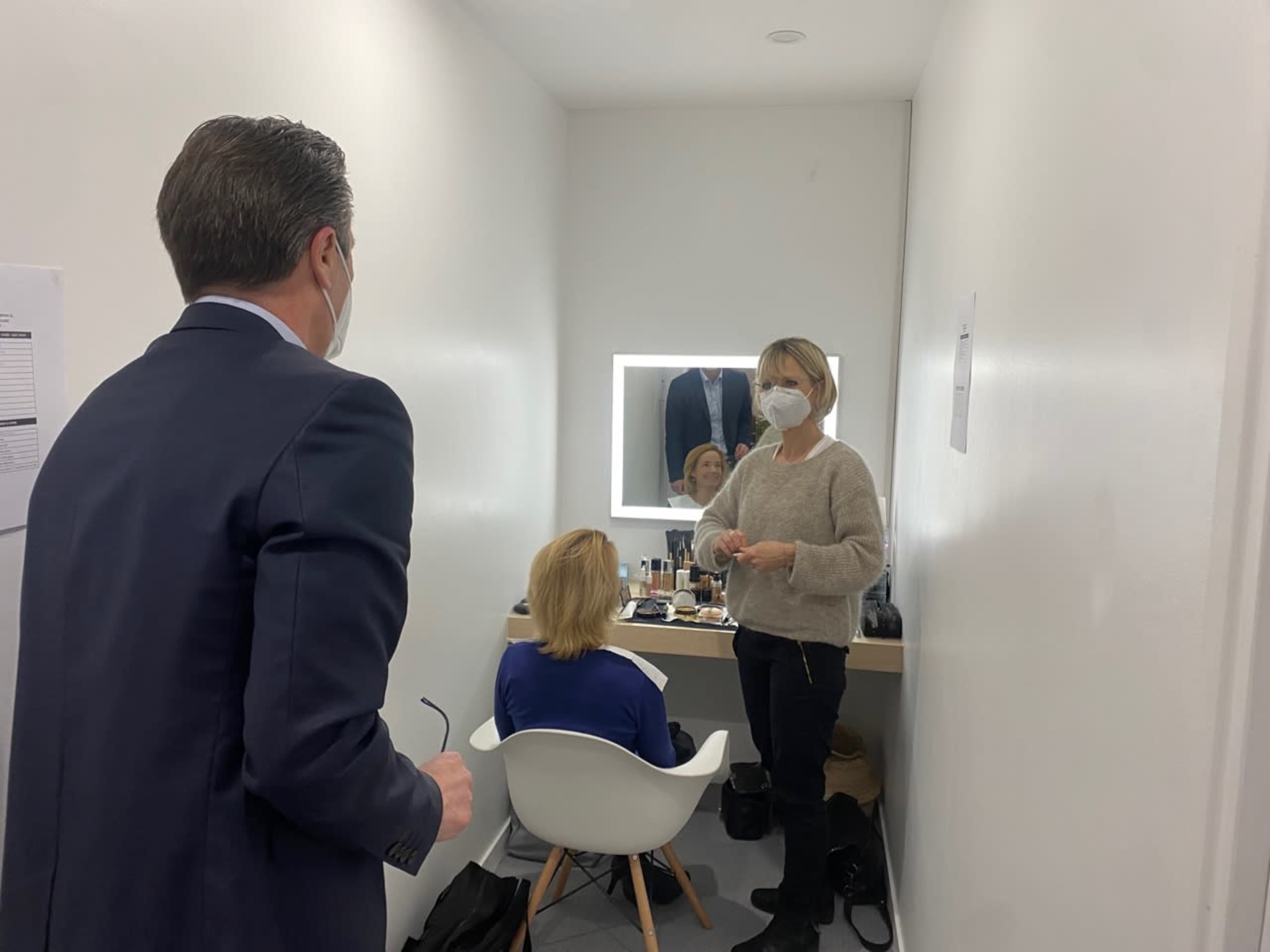 A man in a suit stands talking to a woman applying makeup to another woman seated in front of a mirror in a brightly lit room.