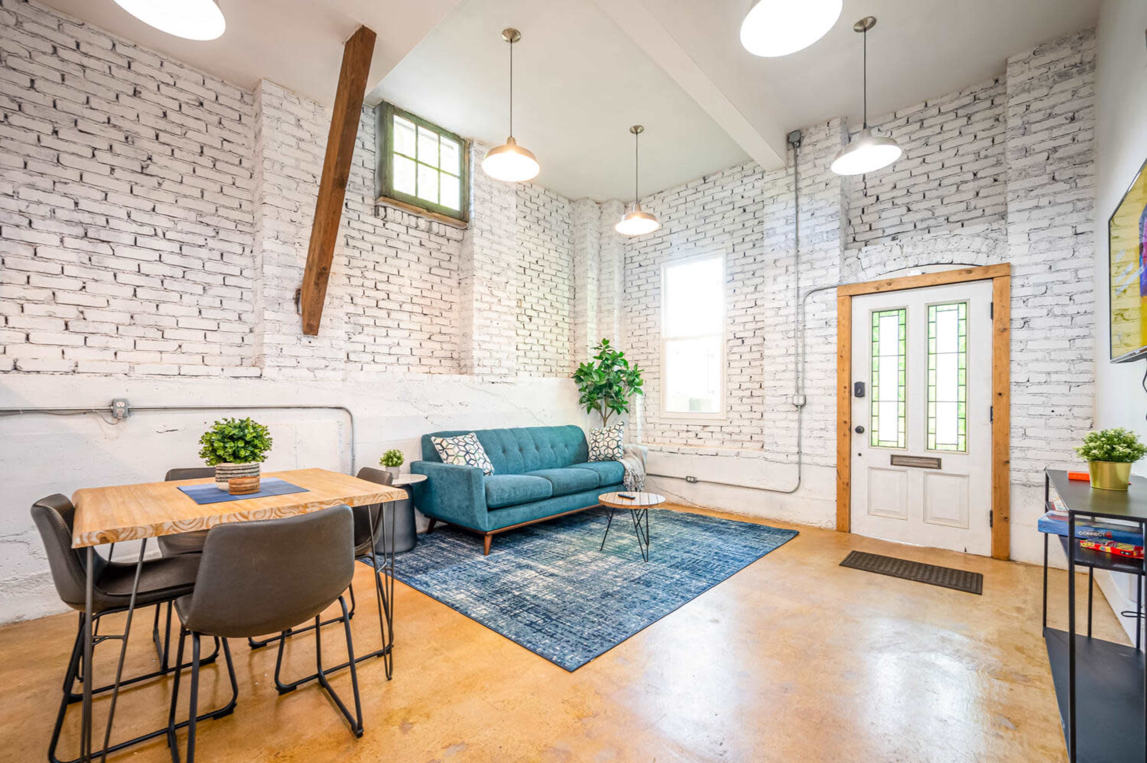 A modern living space featuring a teal couch, a wooden dining table with chairs, and a large area rug on a concrete floor, with white brick walls and large windows allowing natural light.