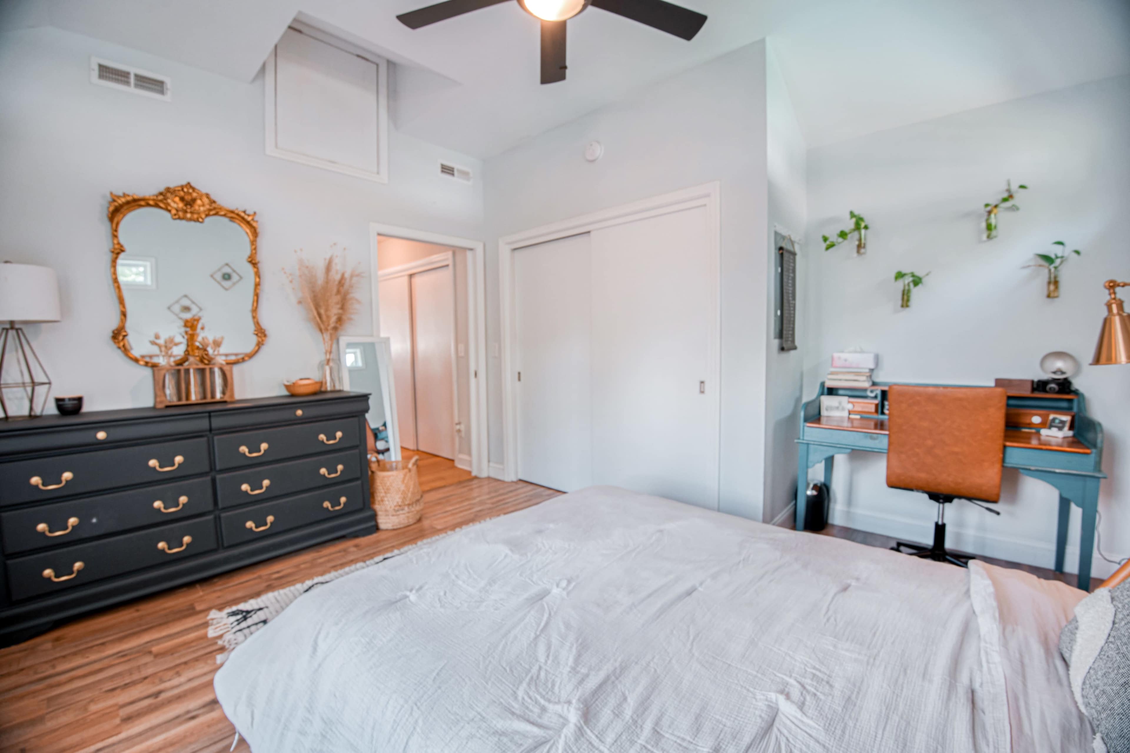A modern bedroom featuring a bed with a light gray cover, a dark dresser with brass handles, a desk with a leather chair, and a few decorative plants on the wall.