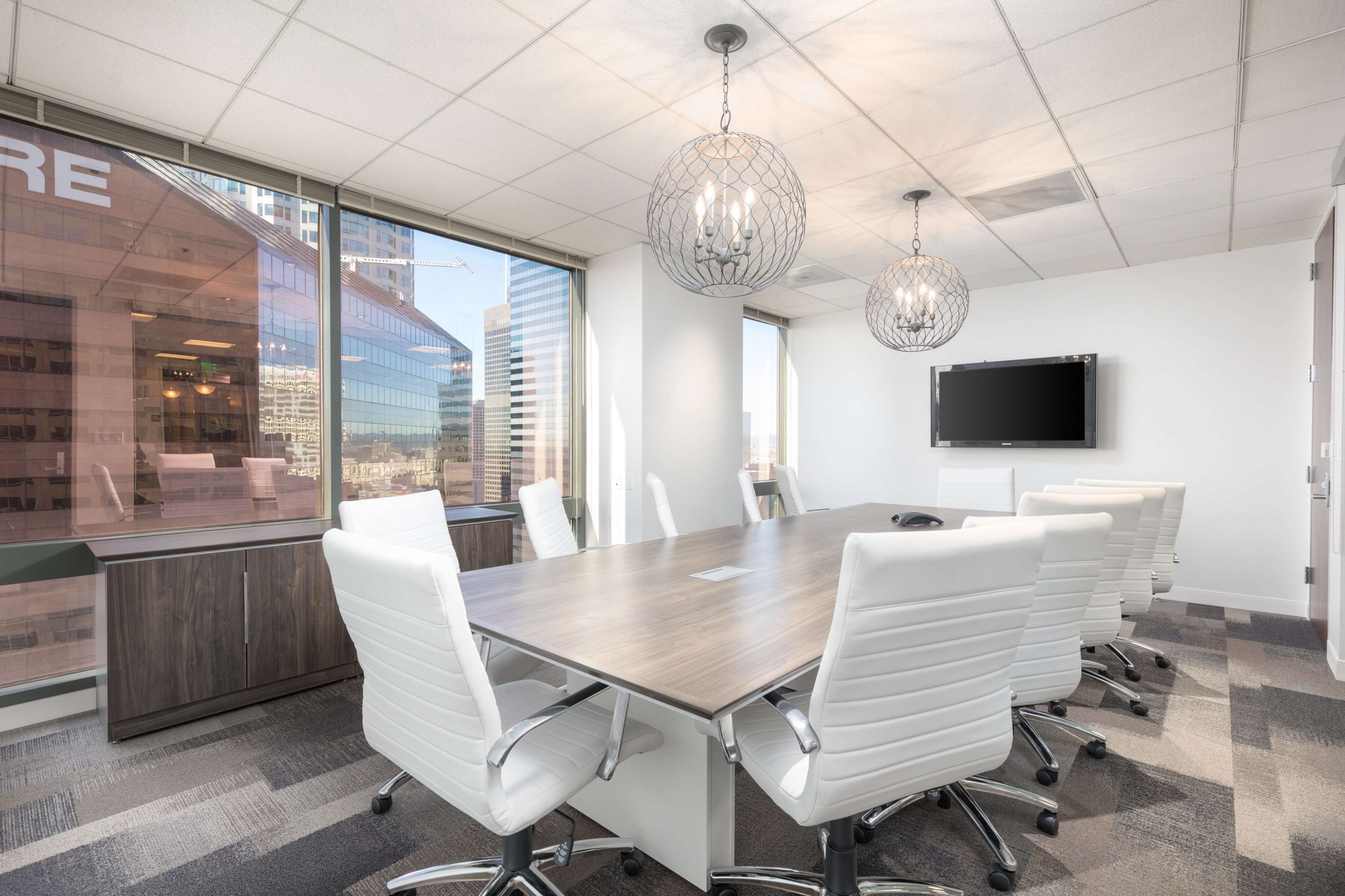 A modern conference room with a large wooden table surrounded by white chairs, large windows revealing a city view, and two pendant lights overhead.