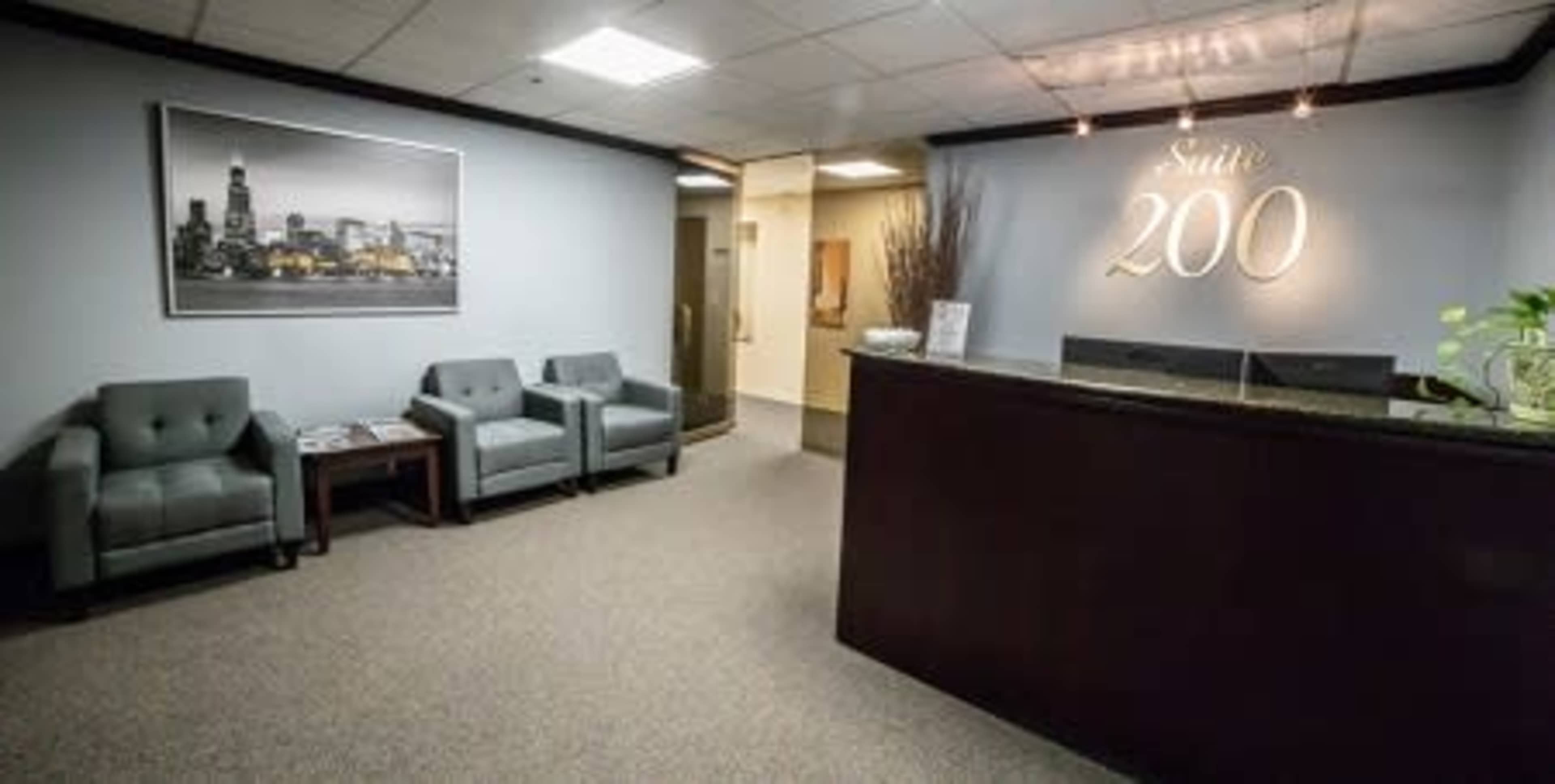 A modern office reception area with gray seating, a front desk labeled "Suite 200," and a large photograph of a city skyline on the wall.