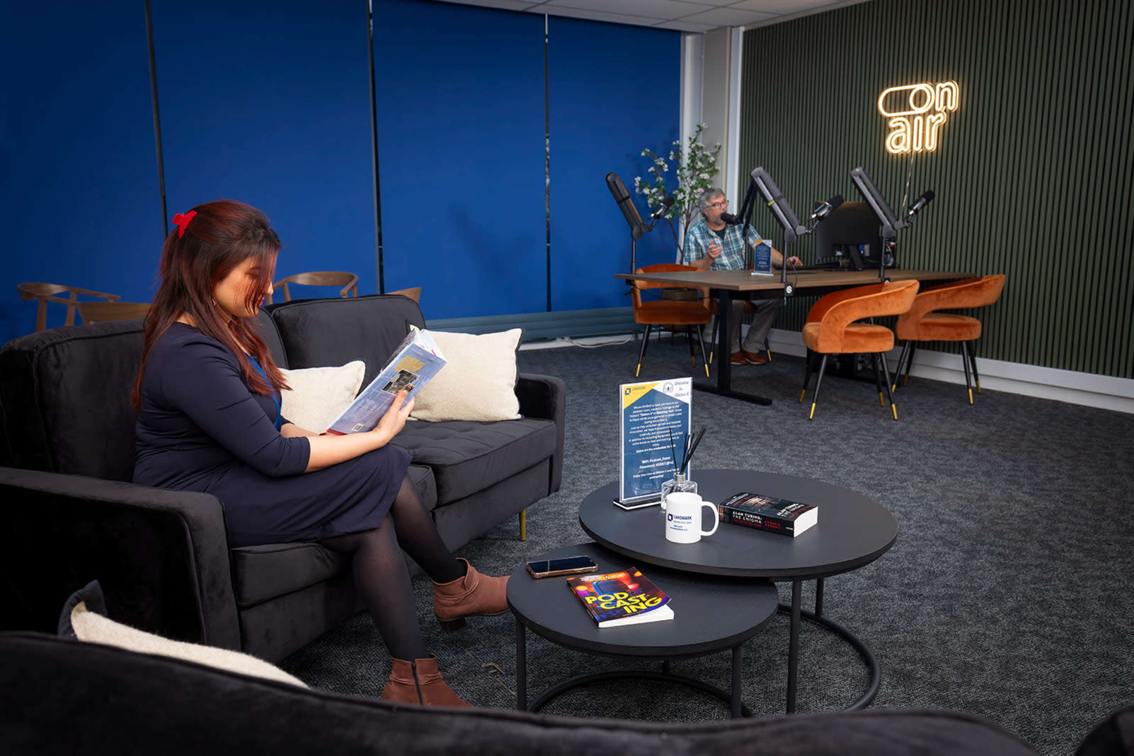 A woman reads a book on a couch in a modern lounge area while a man works at a table in the background.