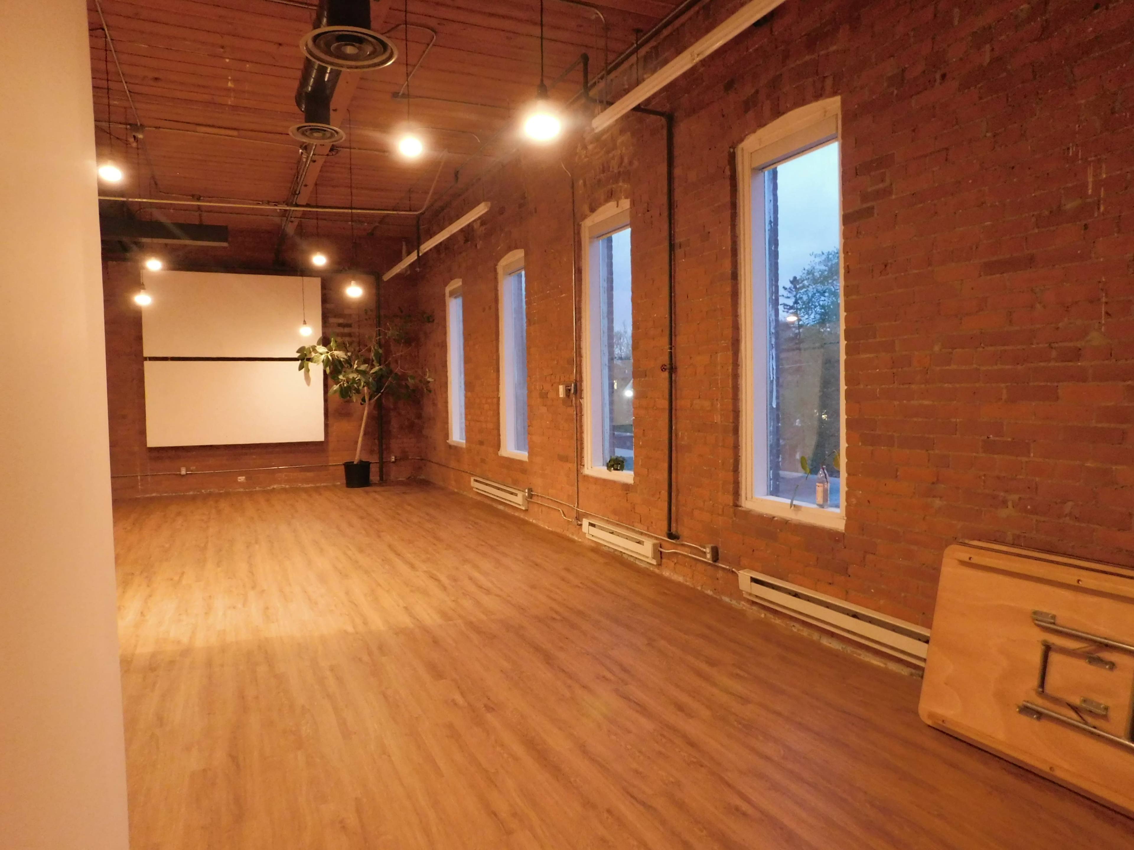 An empty room with exposed brick walls, hardwood flooring, large windows, and a projector screen mounted on the far wall.