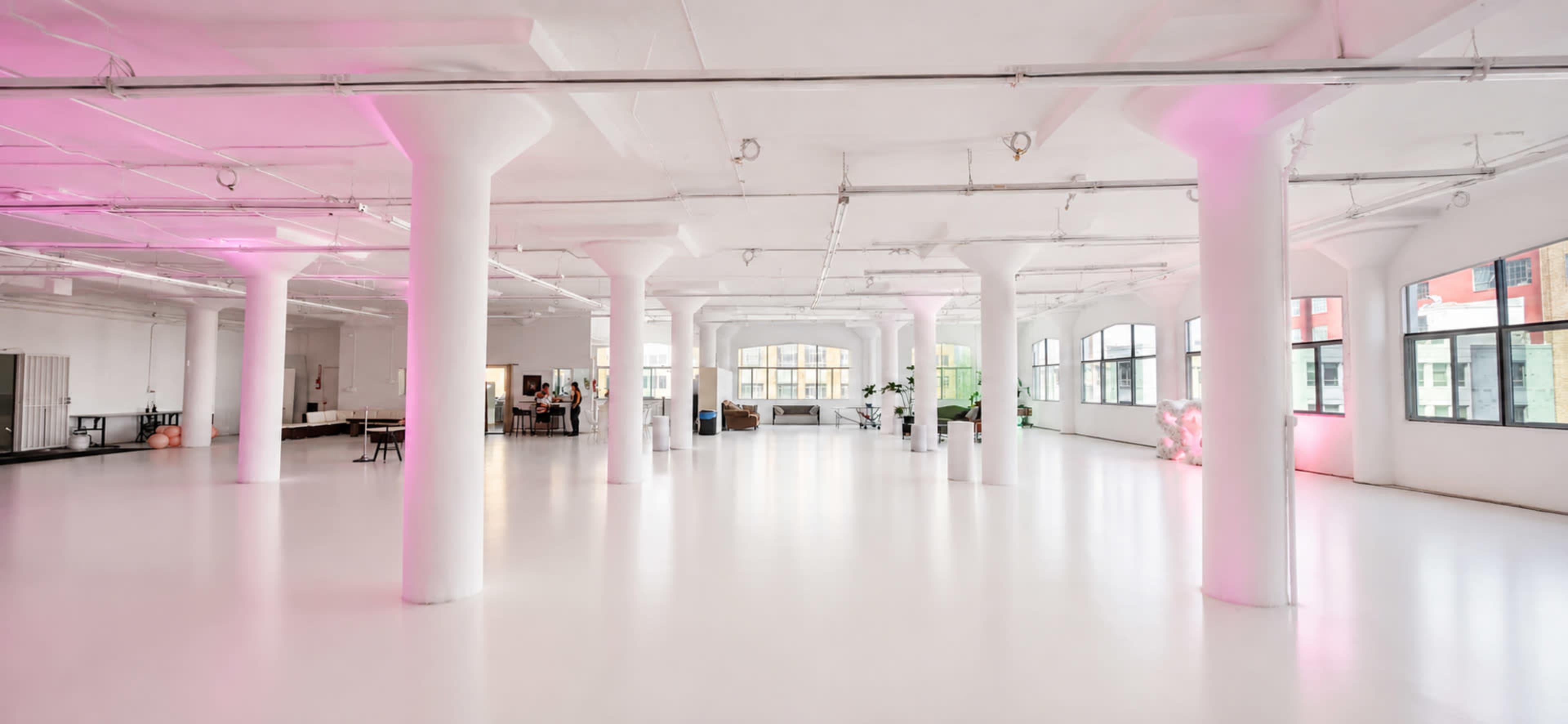 The image shows a spacious, brightly lit interior with white floors, large windows, and pink accent lighting on the columns.