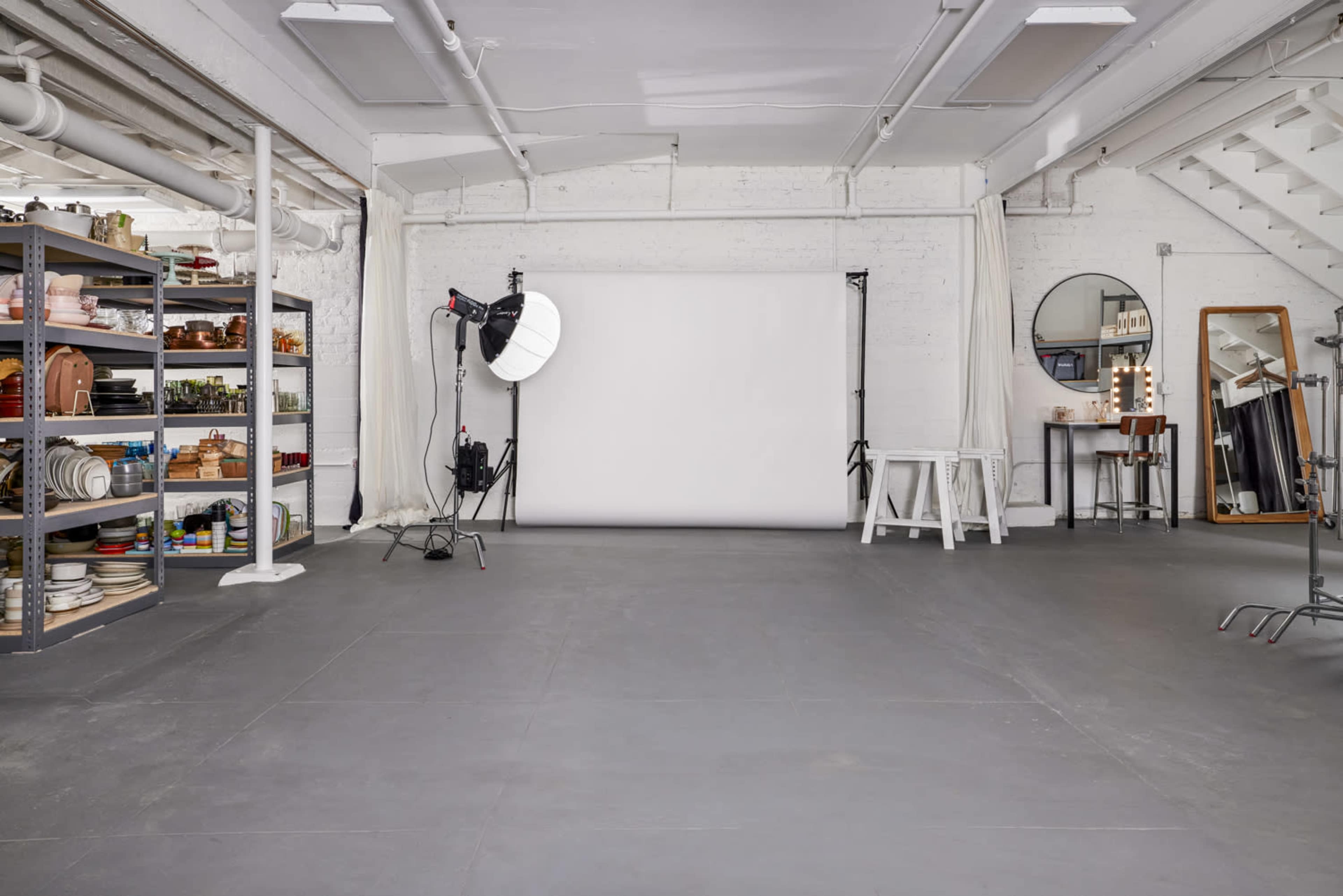 A spacious photography studio with a blank backdrop, lighting equipment, a shelving unit filled with various props, and a large mirror.