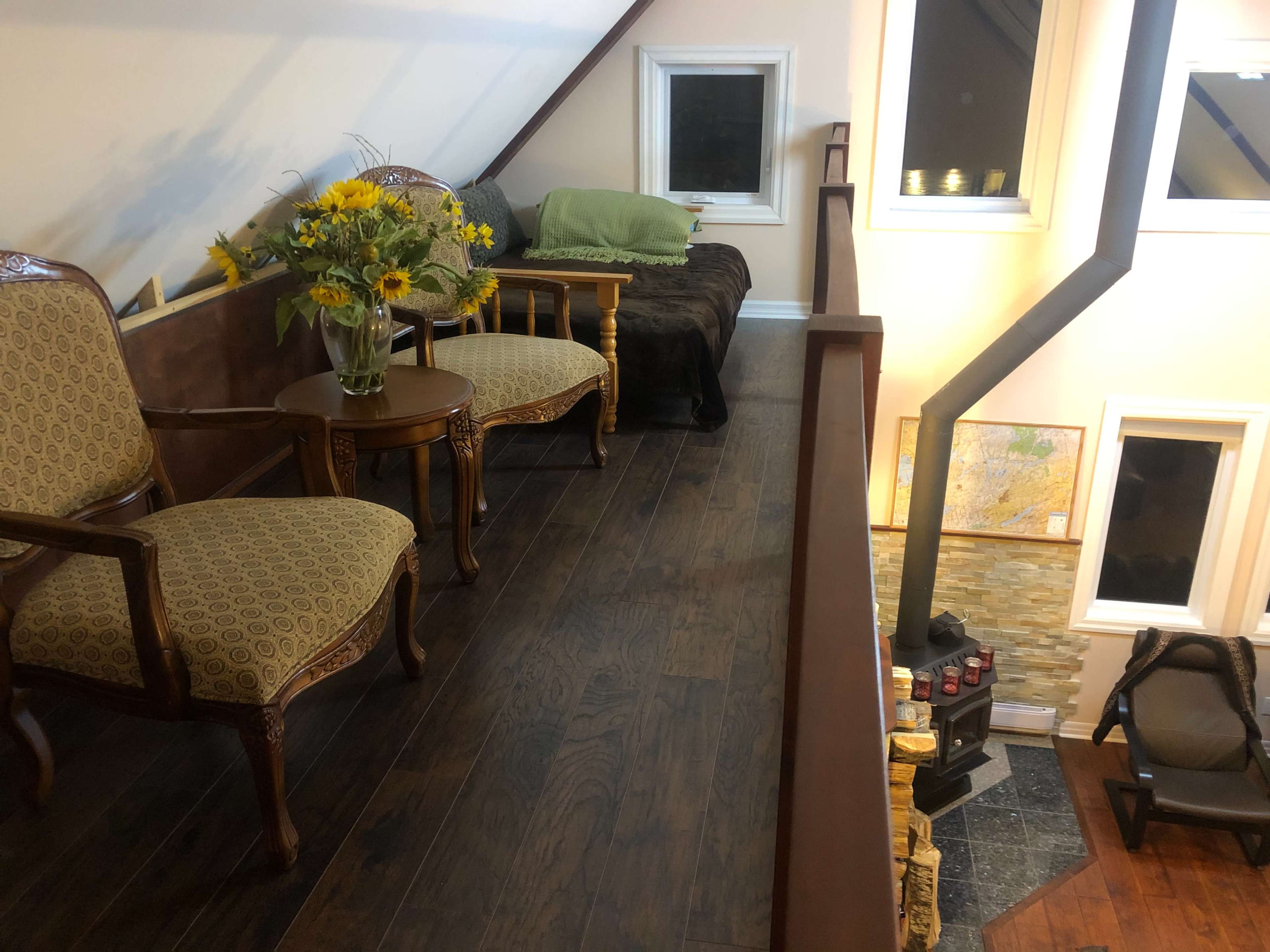 A loft area with two patterned chairs and a small table, decorated with a vase of sunflowers, adjacent to a green couch and a wood-burning stove.