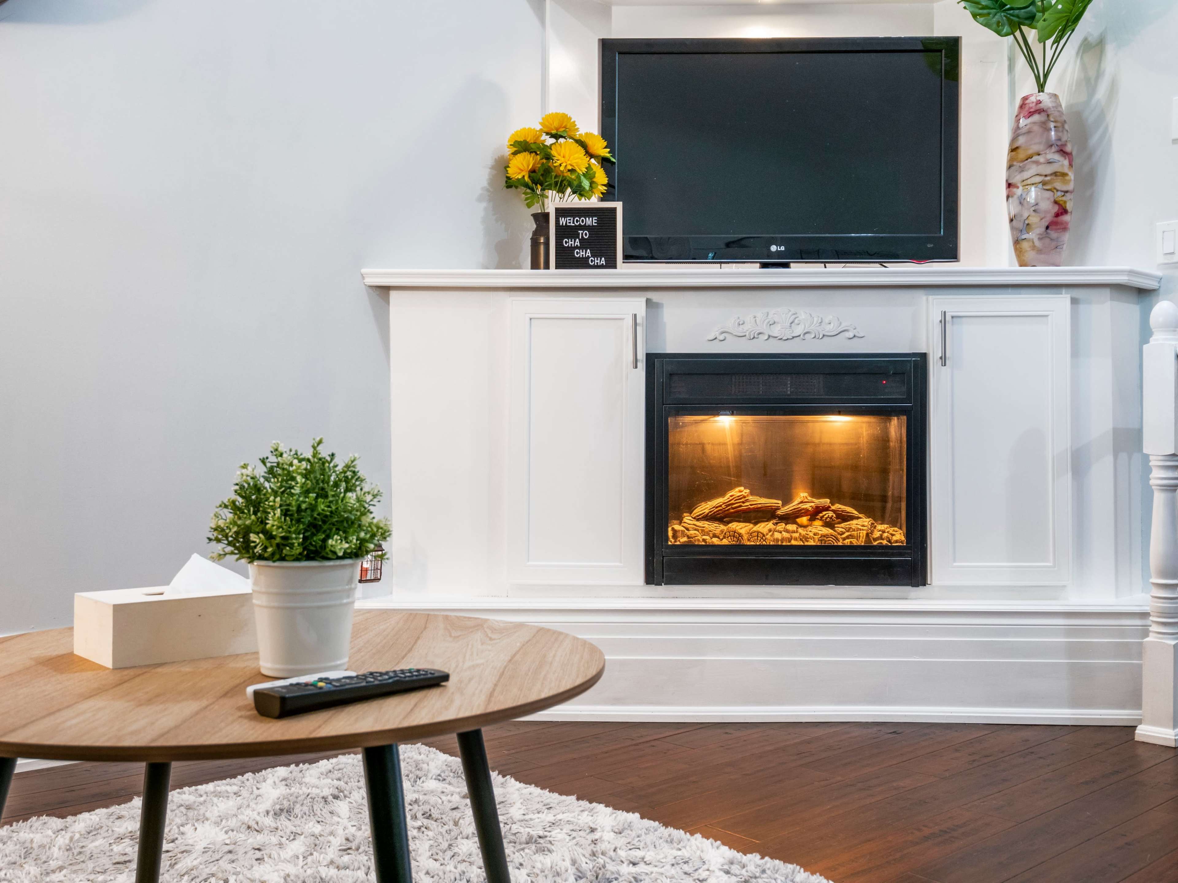 A living room features a modern fireplace beneath a mounted television, with a coffee table and decorative plants.