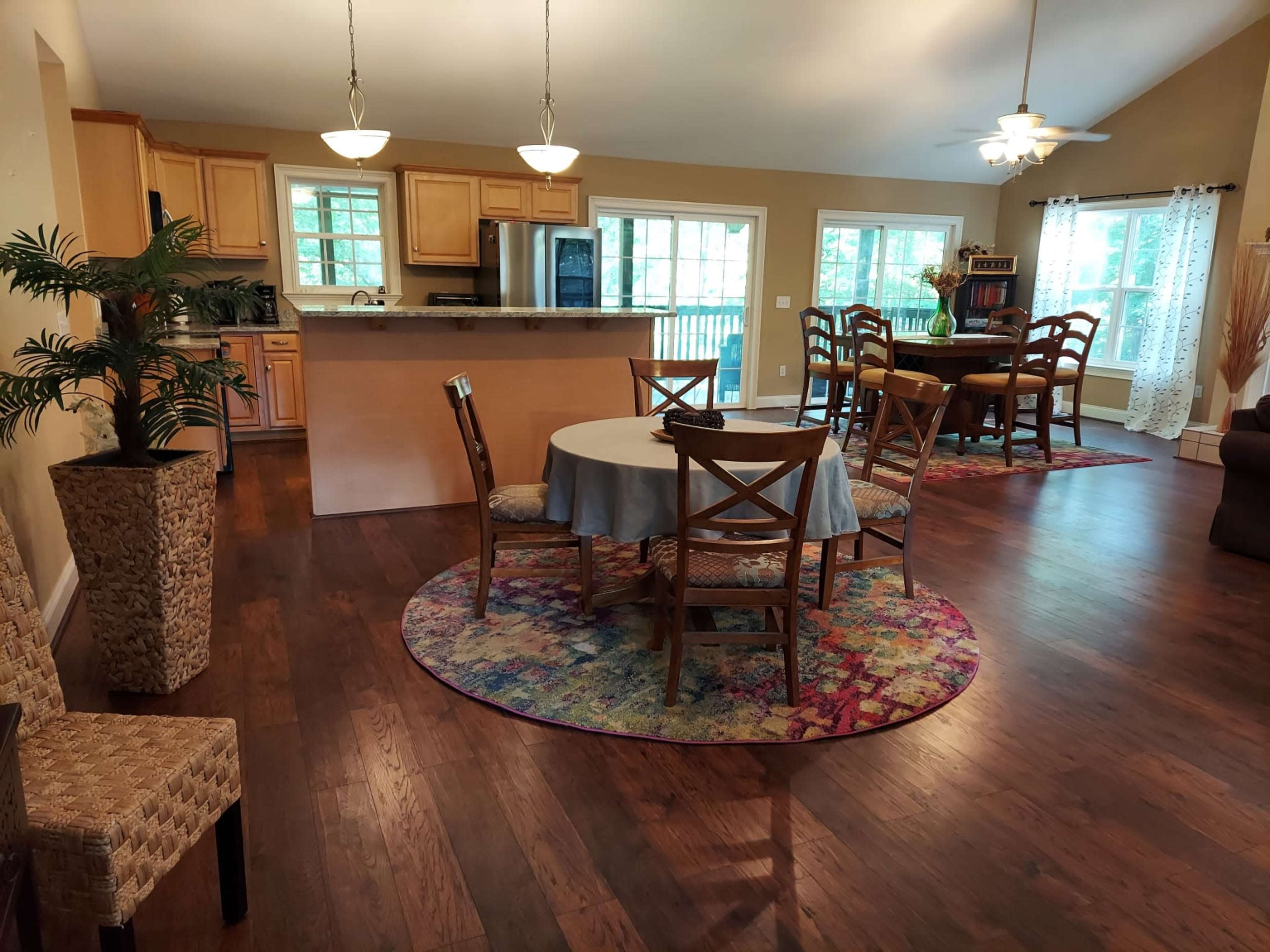 A spacious living area features a dining table surrounded by chairs, with a kitchen in the background and large windows illuminating the space.
