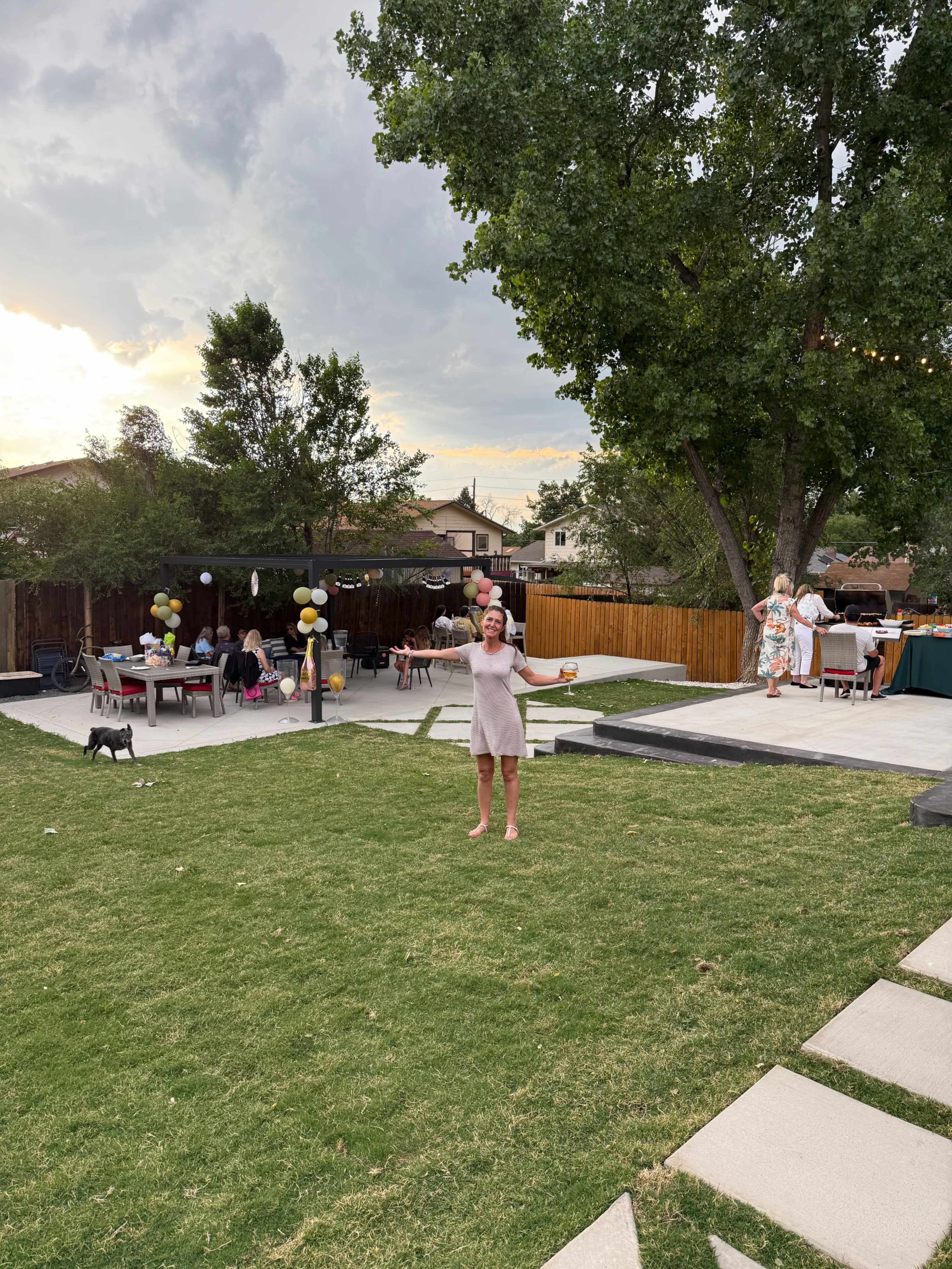 A woman stands in a grassy backyard with seating areas and decorations, while other people mingle in the background.