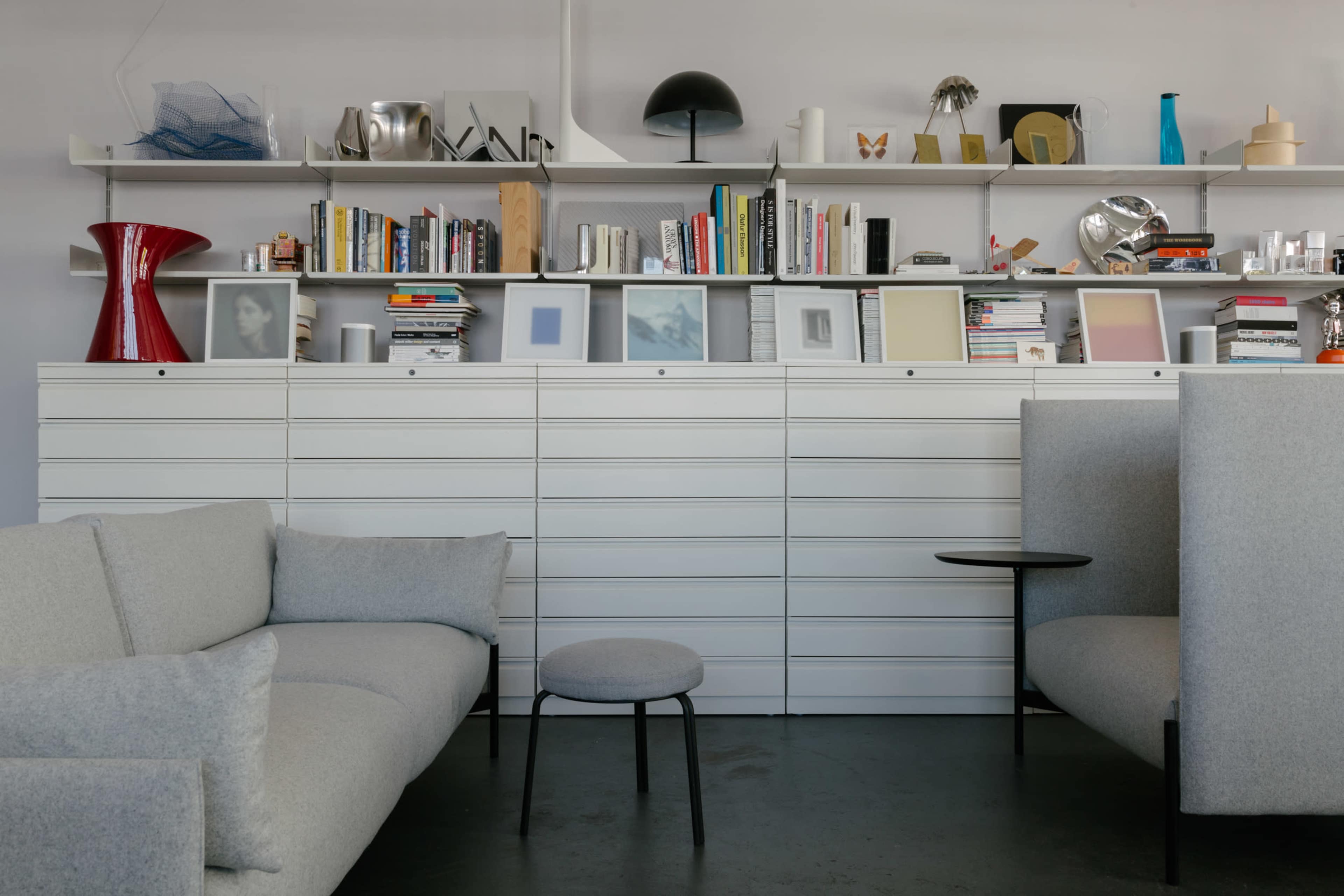 A modern interior with a light gray sofa, a small round stool, and a minimalist storage unit filled with books and decorative objects on a shelf above.