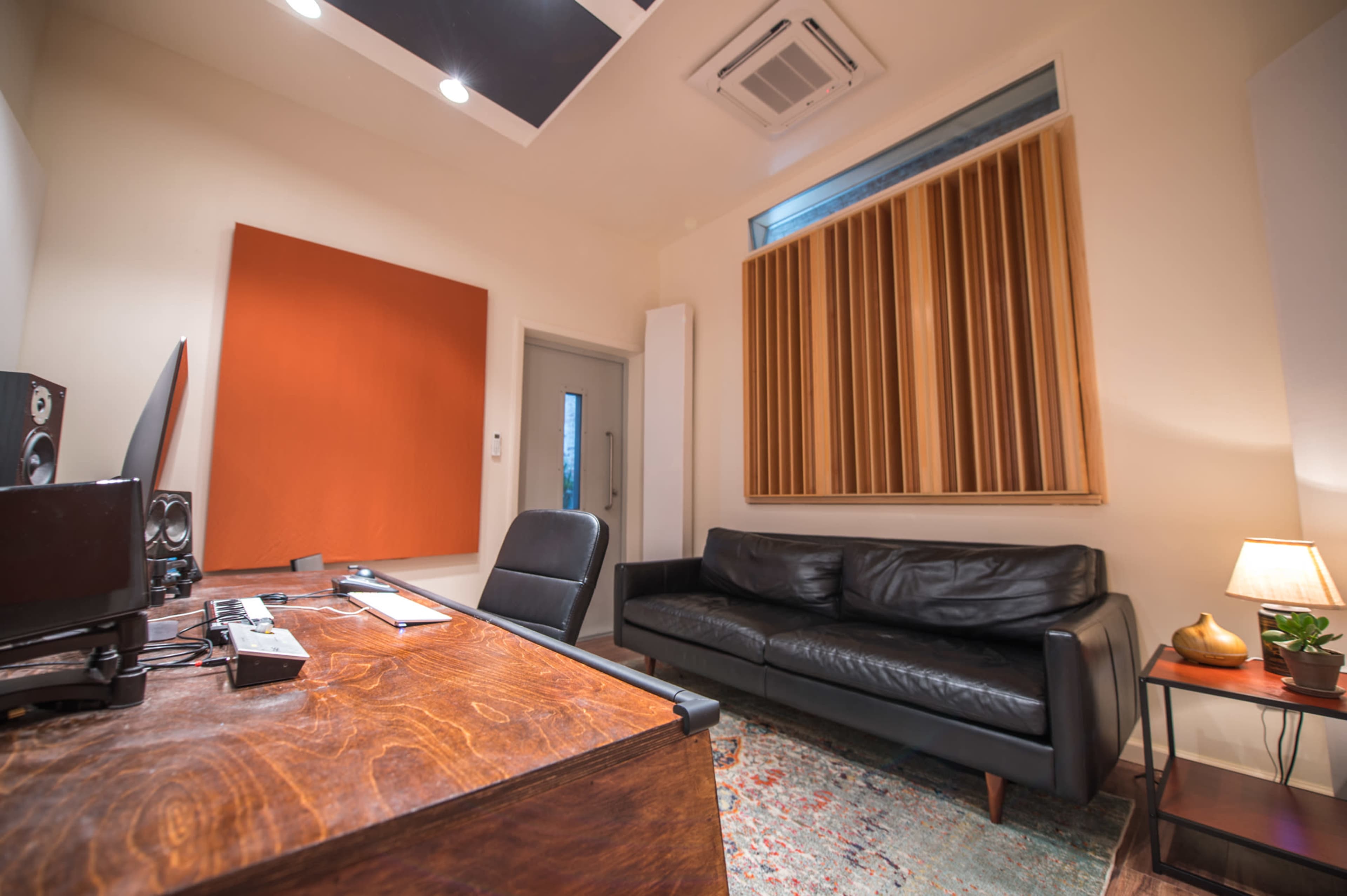 A small music studio features a wooden desk, a black leather sofa, and soundproofing panels on the walls.
